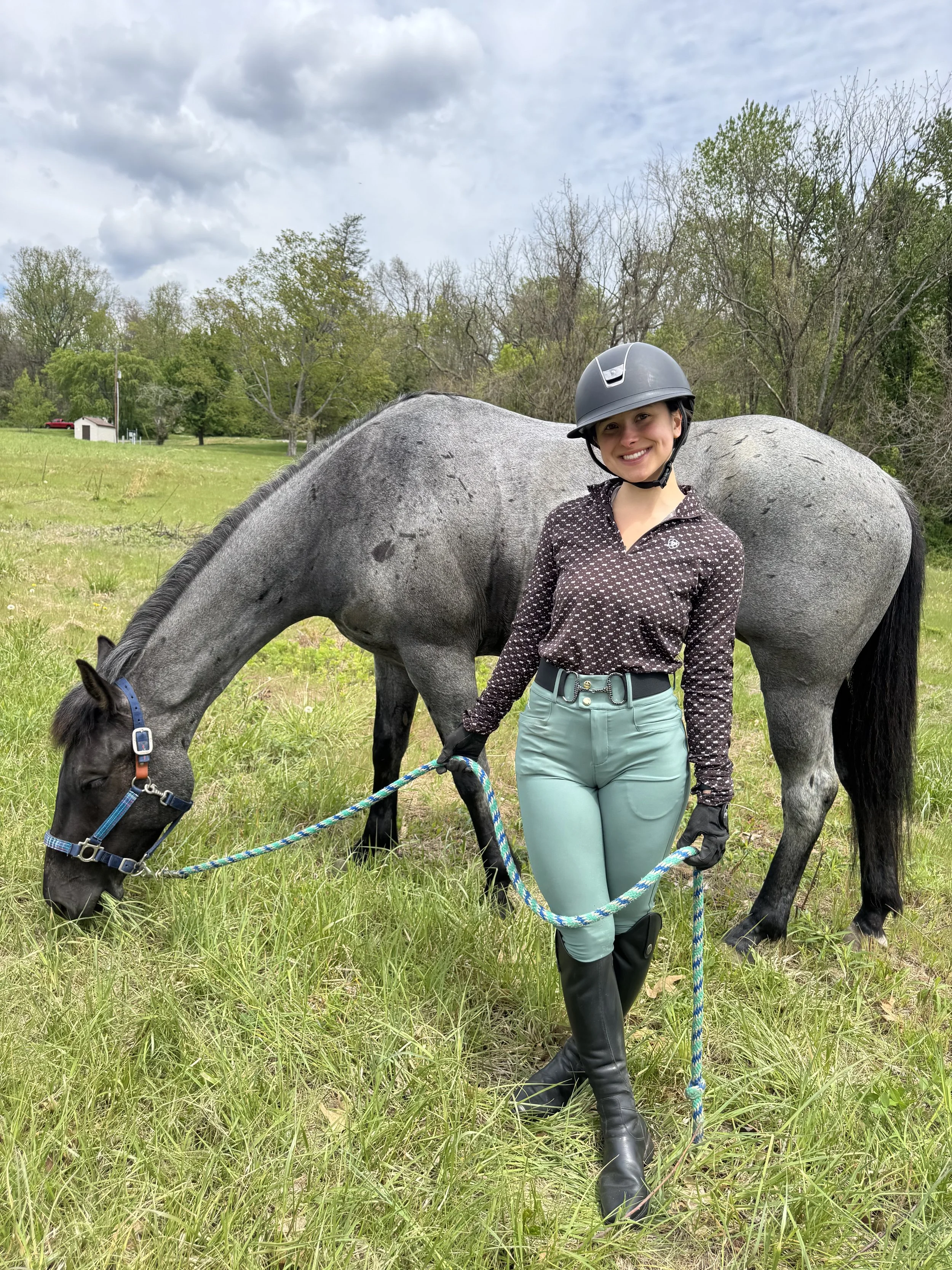A smiling woman wearing riding gear, including a helmet, standing in a grassy field with a gray horse grazing nearby, surrounded by trees and a cloudy sky.
