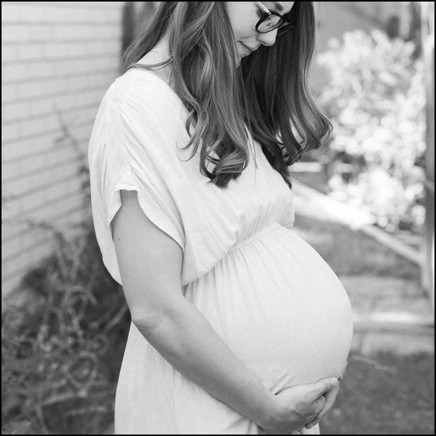 A pregnant woman wearing glasses and a light-colored dress, standing outdoors, gently cradling her baby bump.