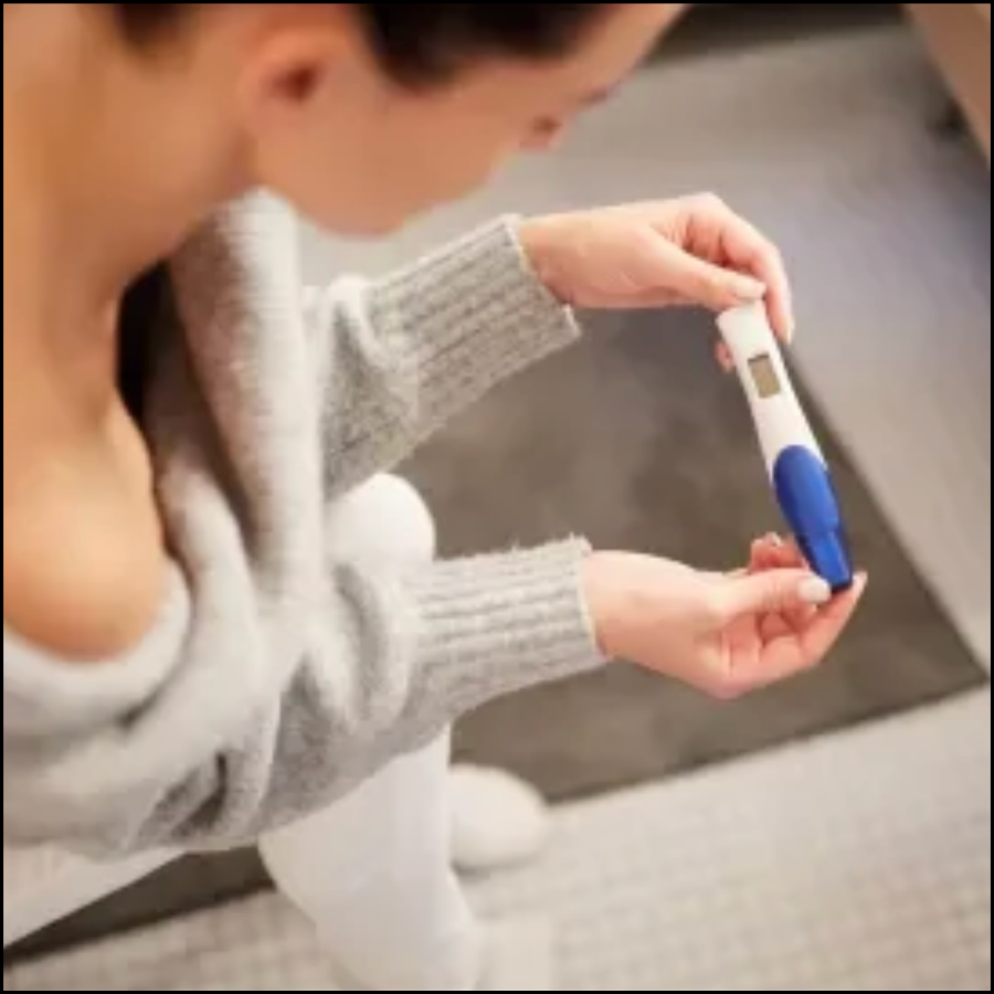 A woman sitting on a couch, wearing a gray sweater and white pants, holding a digital thermometer in her hands, looking at its display.