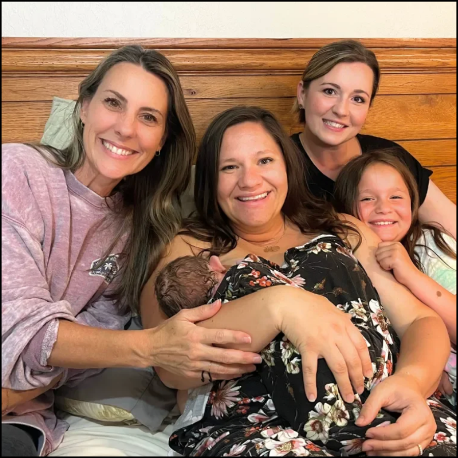 Four women and a young girl sitting on a bed, smiling for a group photo. One woman is holding a newborn baby, possibly their sibling or friend