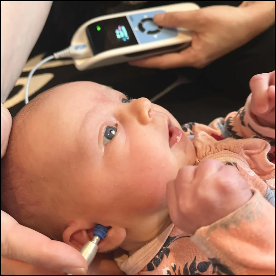 Infant receiving an ear examination with a medical instrument, lying on a caregiver's lap, with an otoscope inserted into her ear, while a healthcare worker adjusts a device in the background.