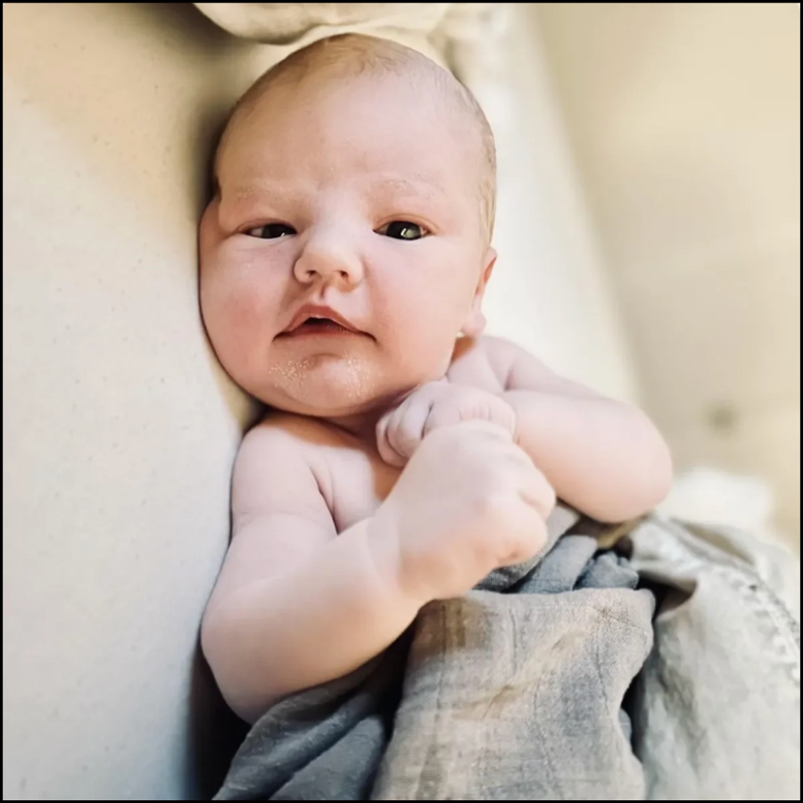 Close-up of a baby lying on a soft surface, looking at the camera with a relaxed expression, hands near face, wearing a cloth diaper.