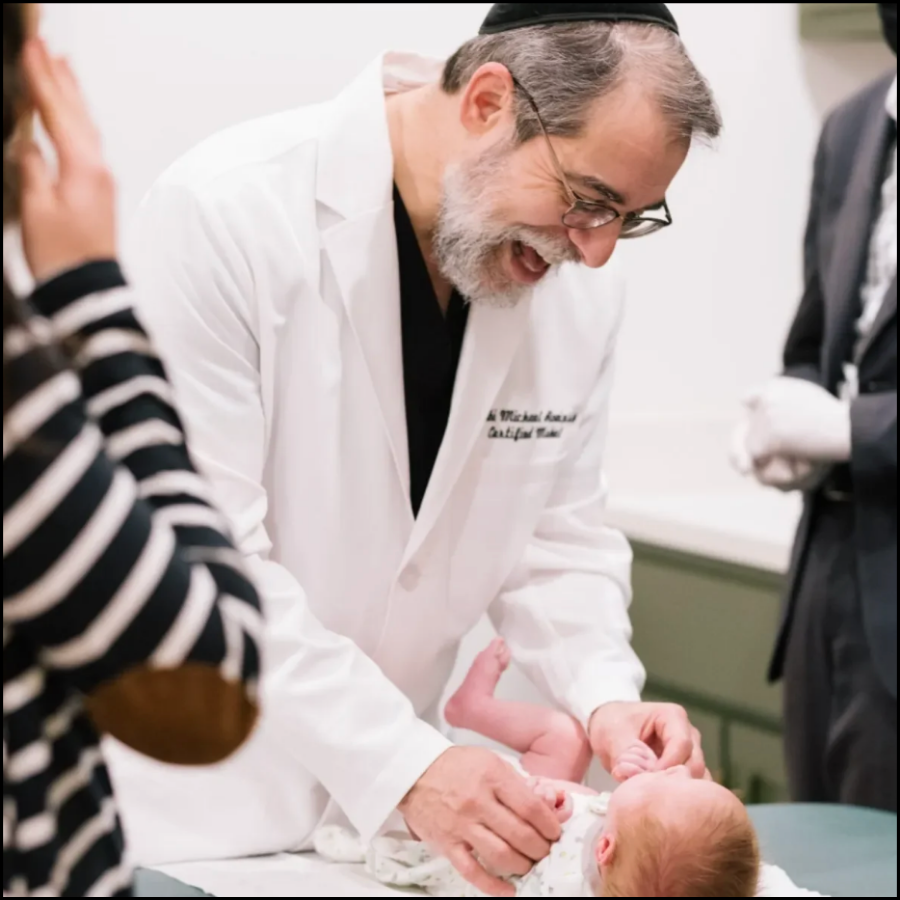 A doctor with glasses and a gray beard, wearing a white lab coat, is smiling as he cares for a newborn baby lying on an examination table. Other people are present, one with a striped shirt.