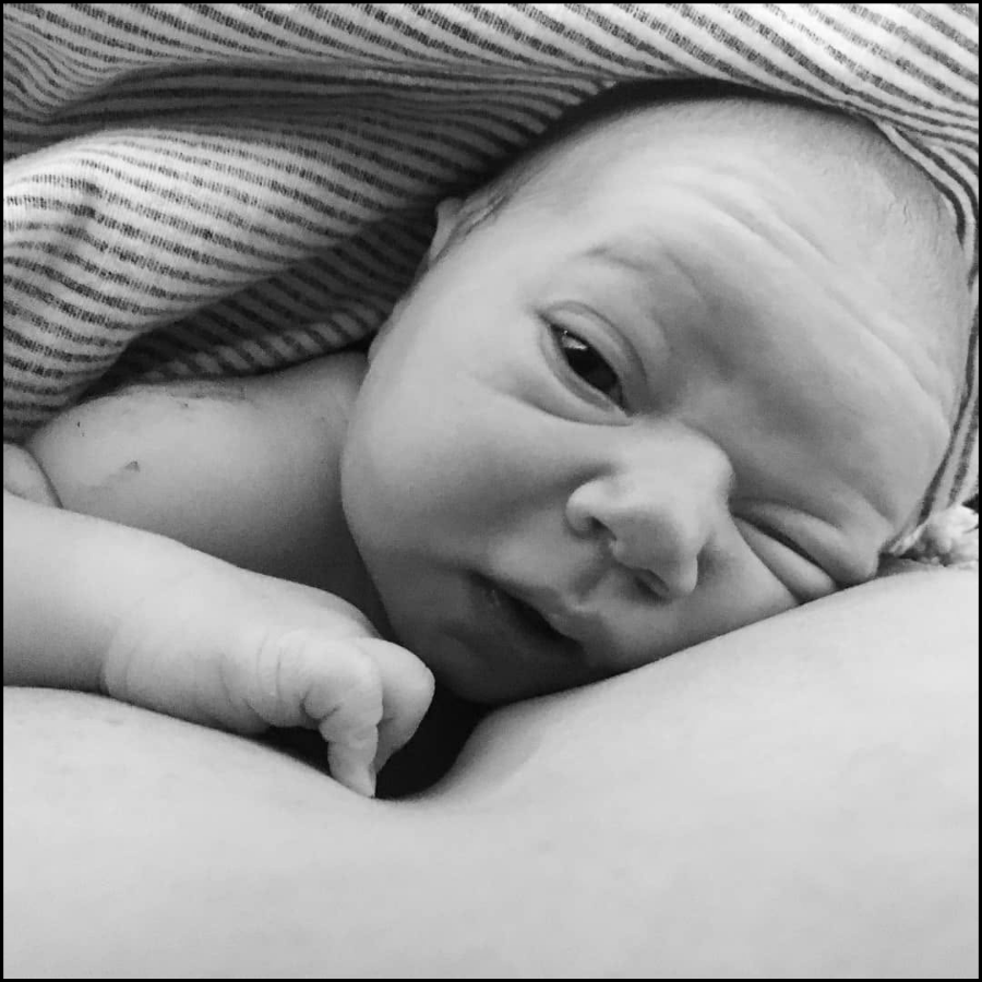 Black and white photo of a newborn baby lying on a bed, partially covered with a striped blanket, with the baby's head resting on a person's arm.