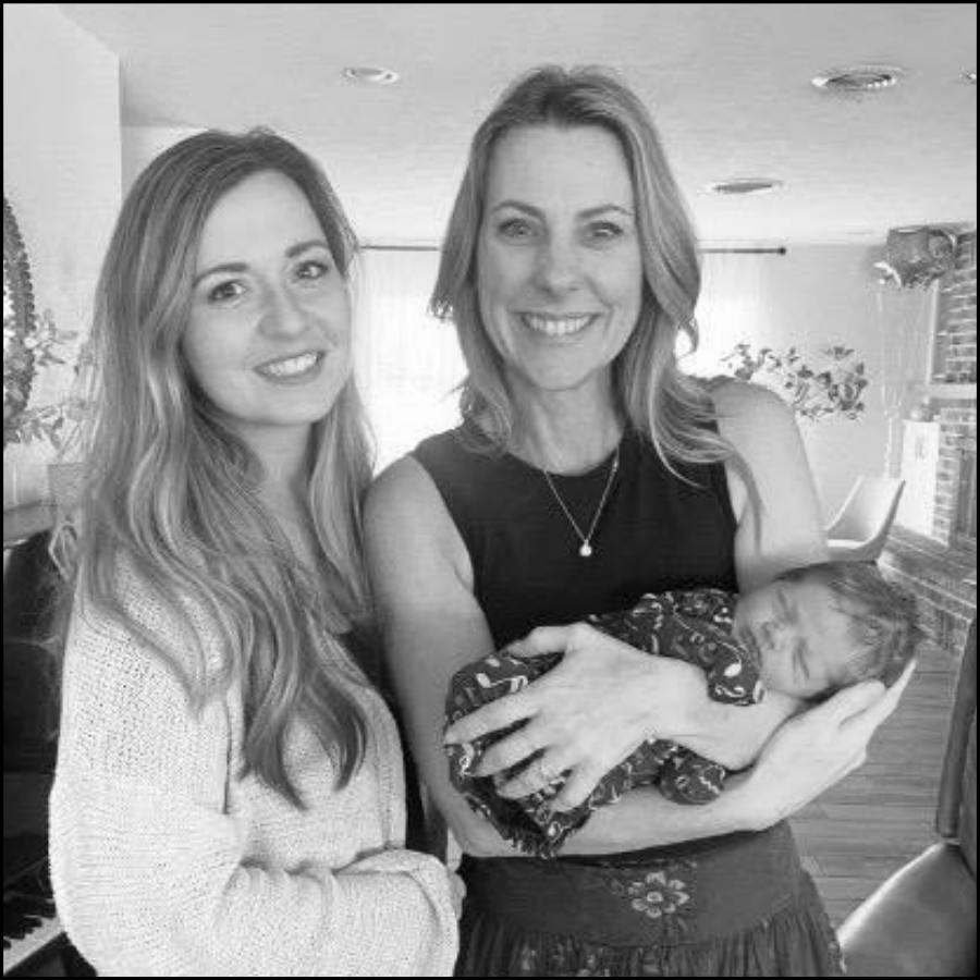 Two women smiling and holding a newborn baby in a home interior with brick wall and plant background.