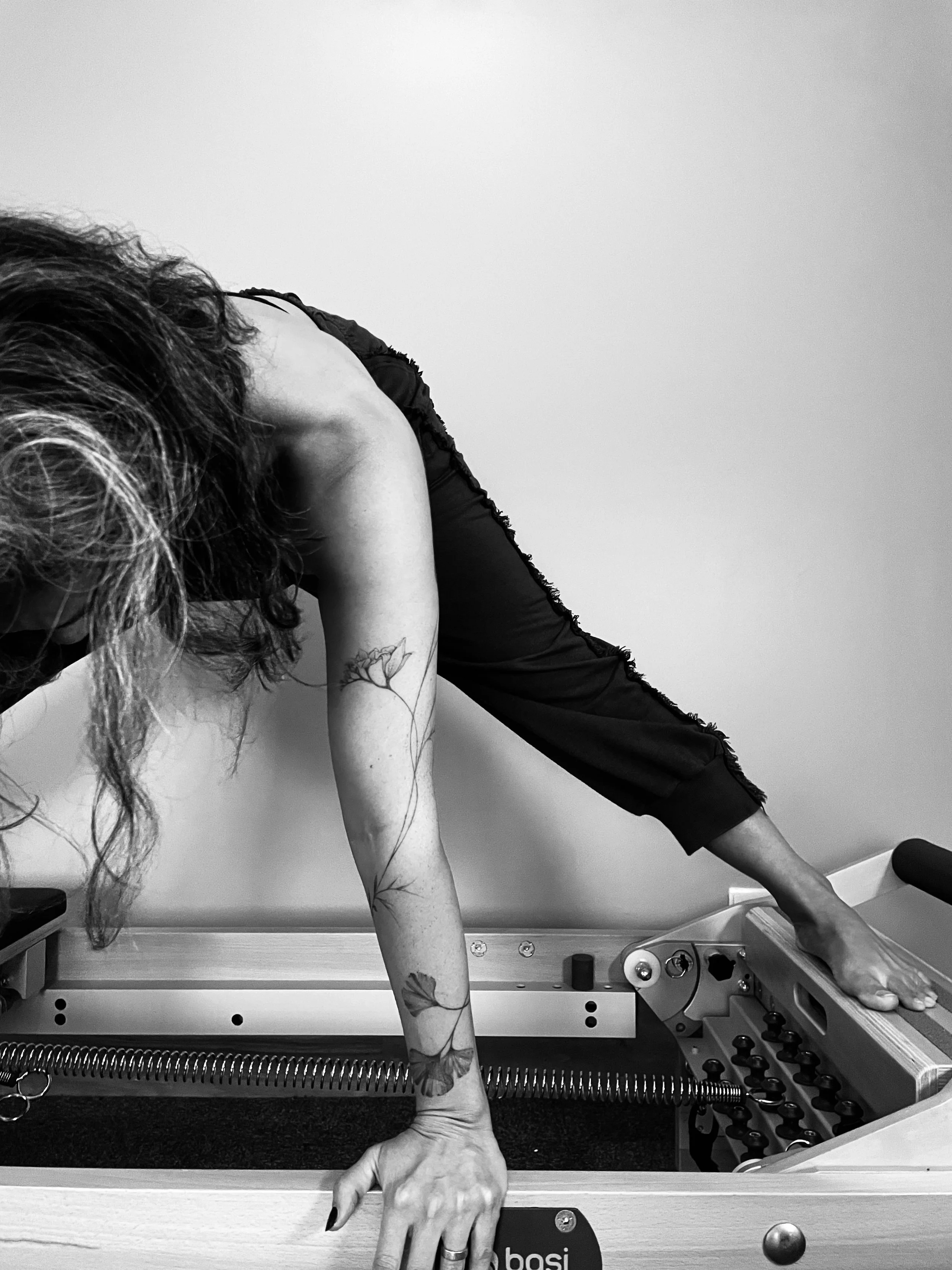 Black-and-white photo of a person practicing yoga on a foam roller, with their leg extended and arm reaching downward, tattoos visible on the arm, in a room with plain wall and foam roller.