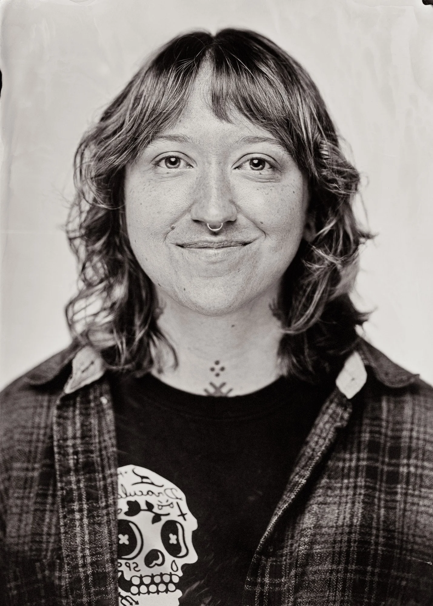 Portrait en noir et blanc d'une femme avec des cheveux ondulés, portant un pull en tartan et un t-shirt noir avec un skull.