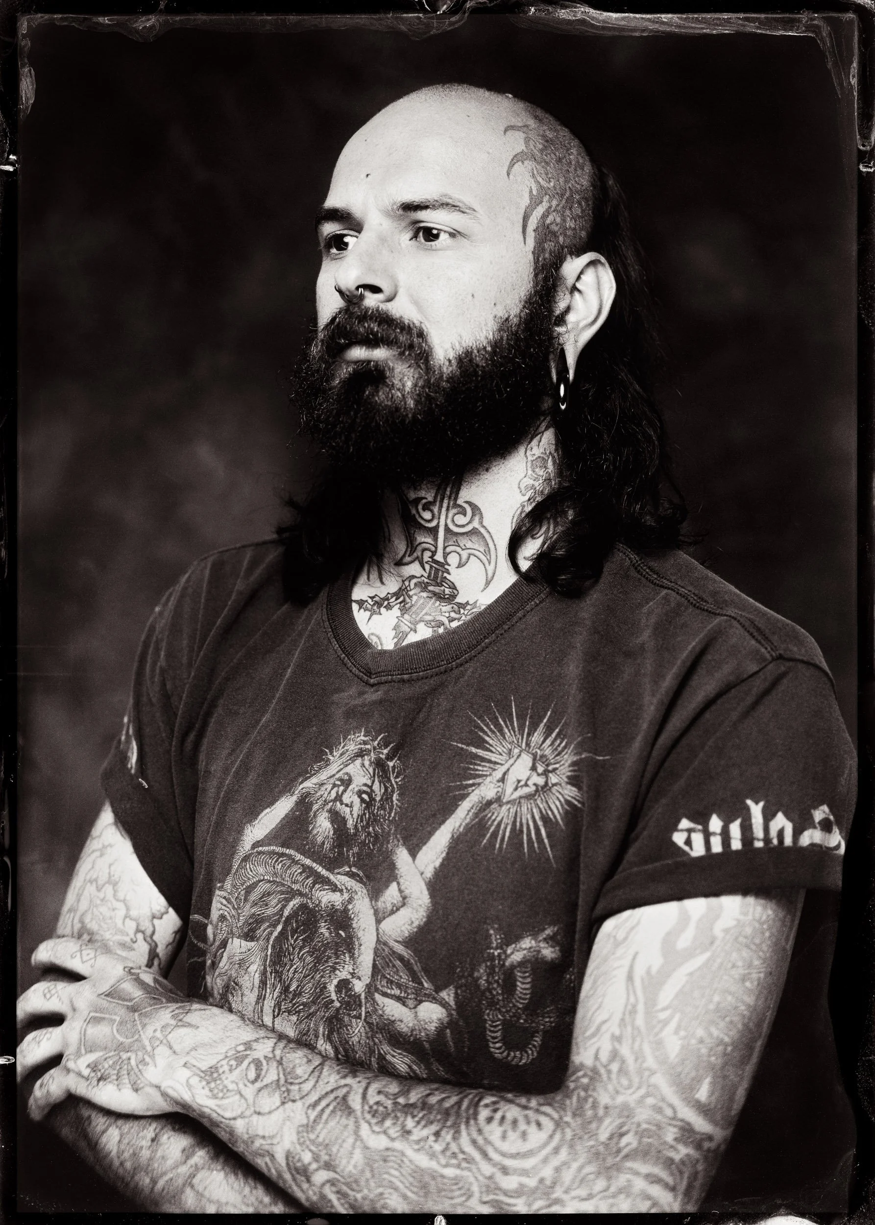 Un homme avec une barbe, des tatouages visibles sur la main et le cou, portant une chemise à carreaux.