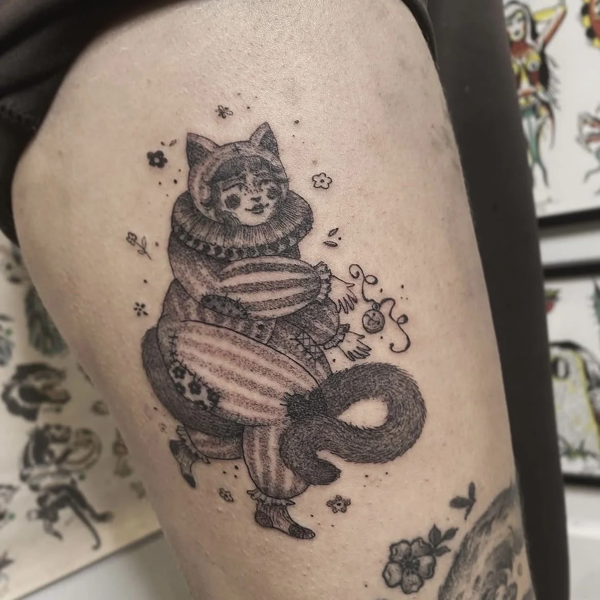 Tatouage d'un personnage avec une tête de chat, habillé comme un clown, tenant une boule rayée, entouré de fleurs et de petits détails.