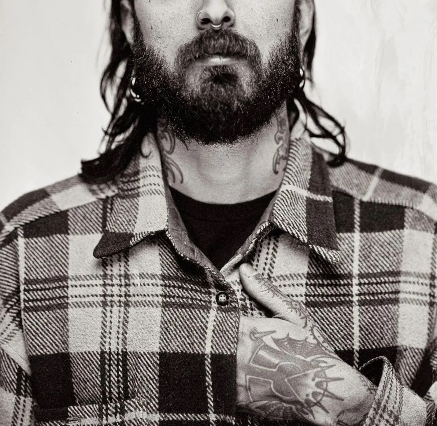 Un homme avec une barbe, des tatouages visibles sur la main et le cou, portant une chemise à carreaux.