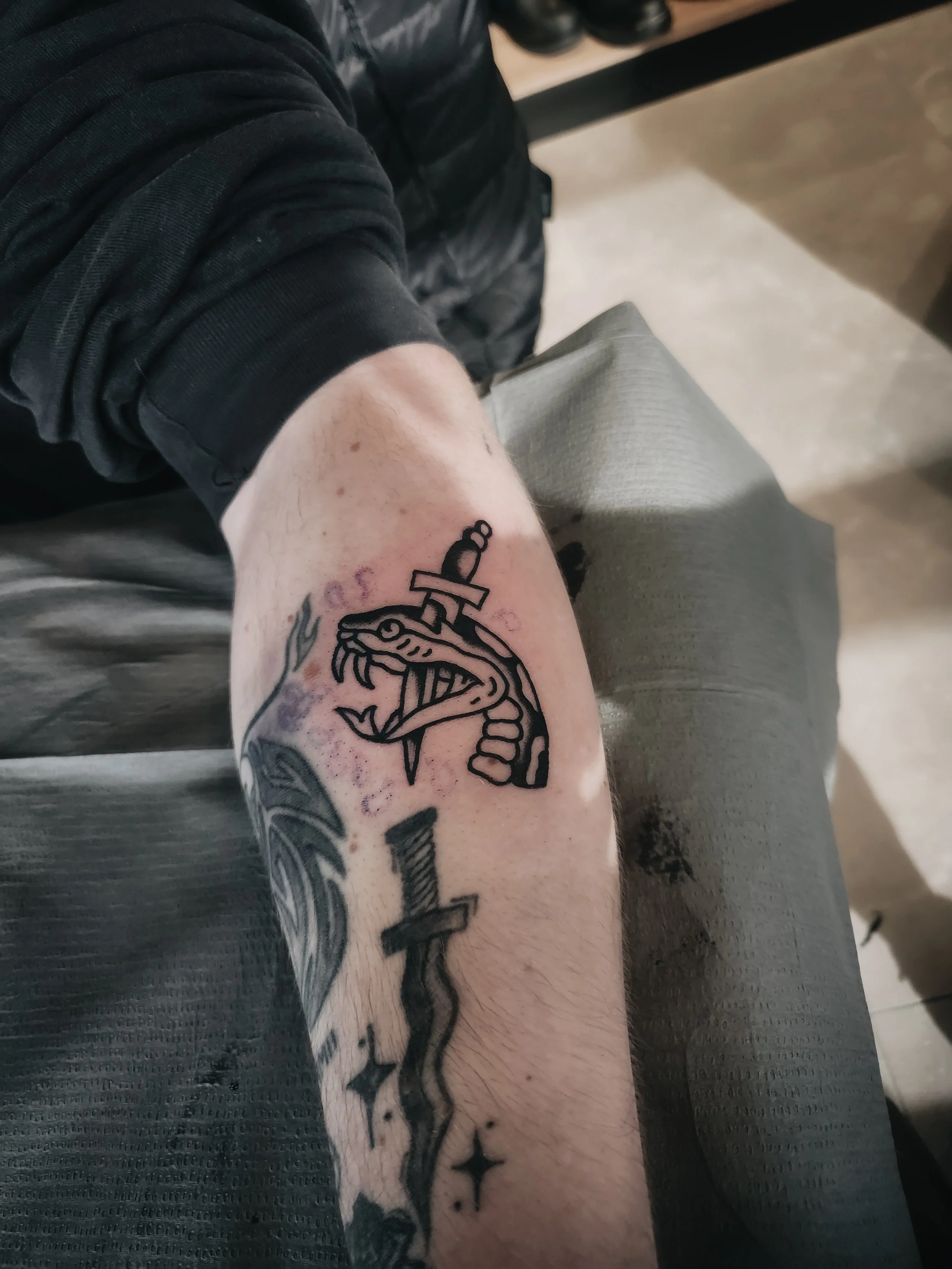 Bras tatoué avec un dessin d'une ancre, un serpent et d'autres éléments noirs, sur le bras d'une personne. La personne porte un vêtement noir à manches longues.