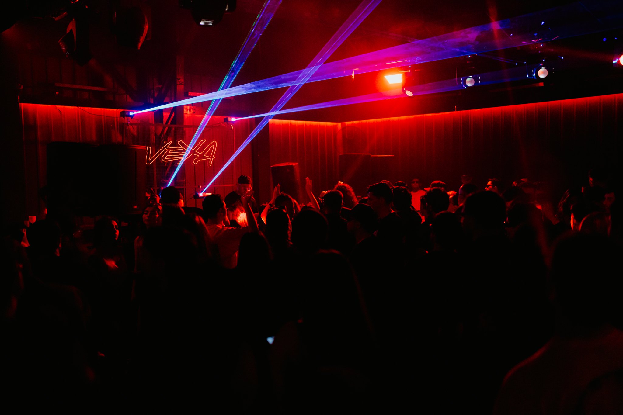 A crowded dance club or concert venue with people dancing in front of a stage, illuminated by colorful laser lights and red lighting, with the word 'VETA' displayed on a neon sign in the background.