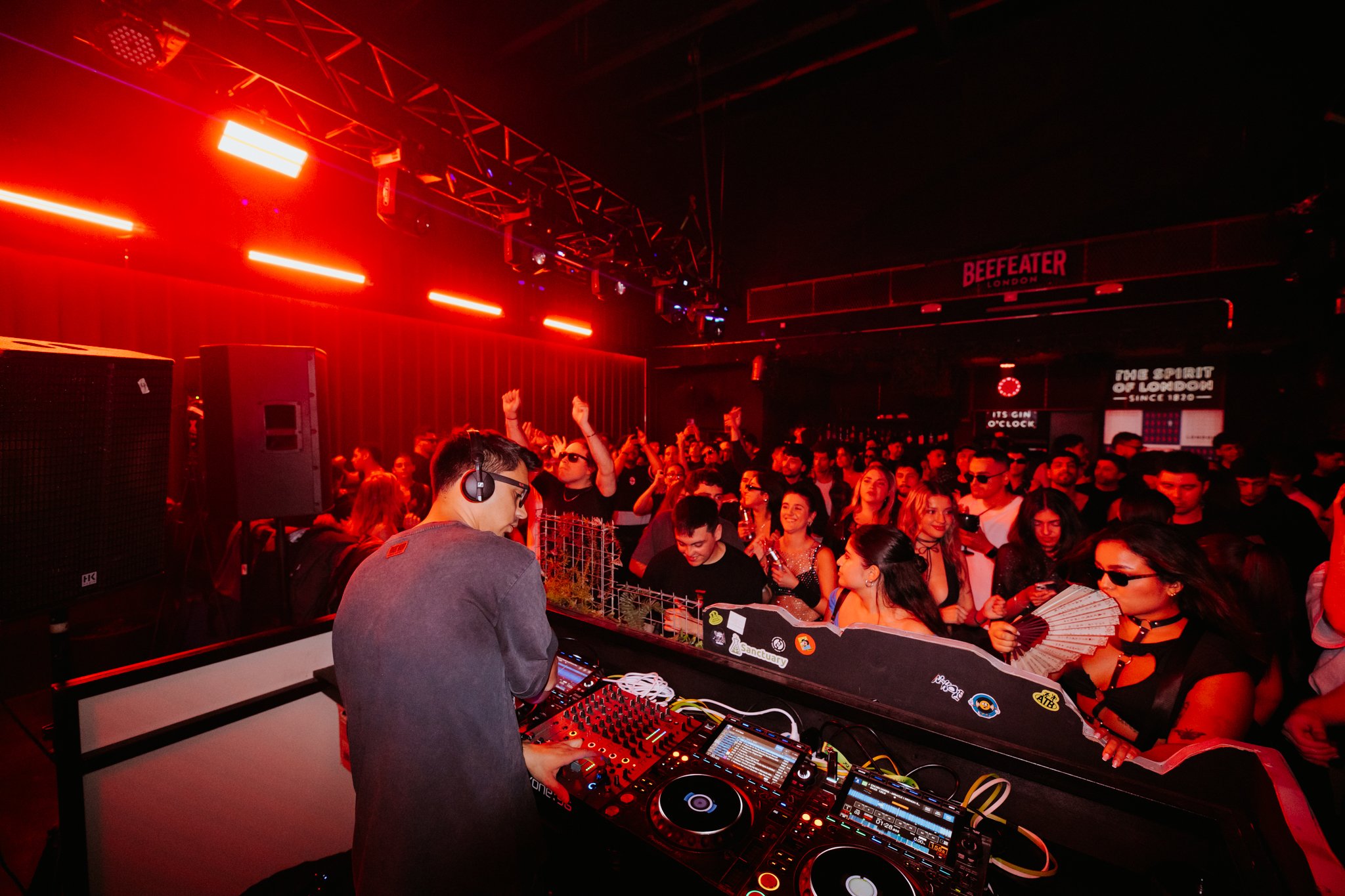 A DJ wearing headphones performs at a nightclub with a crowd dancing under red lights.