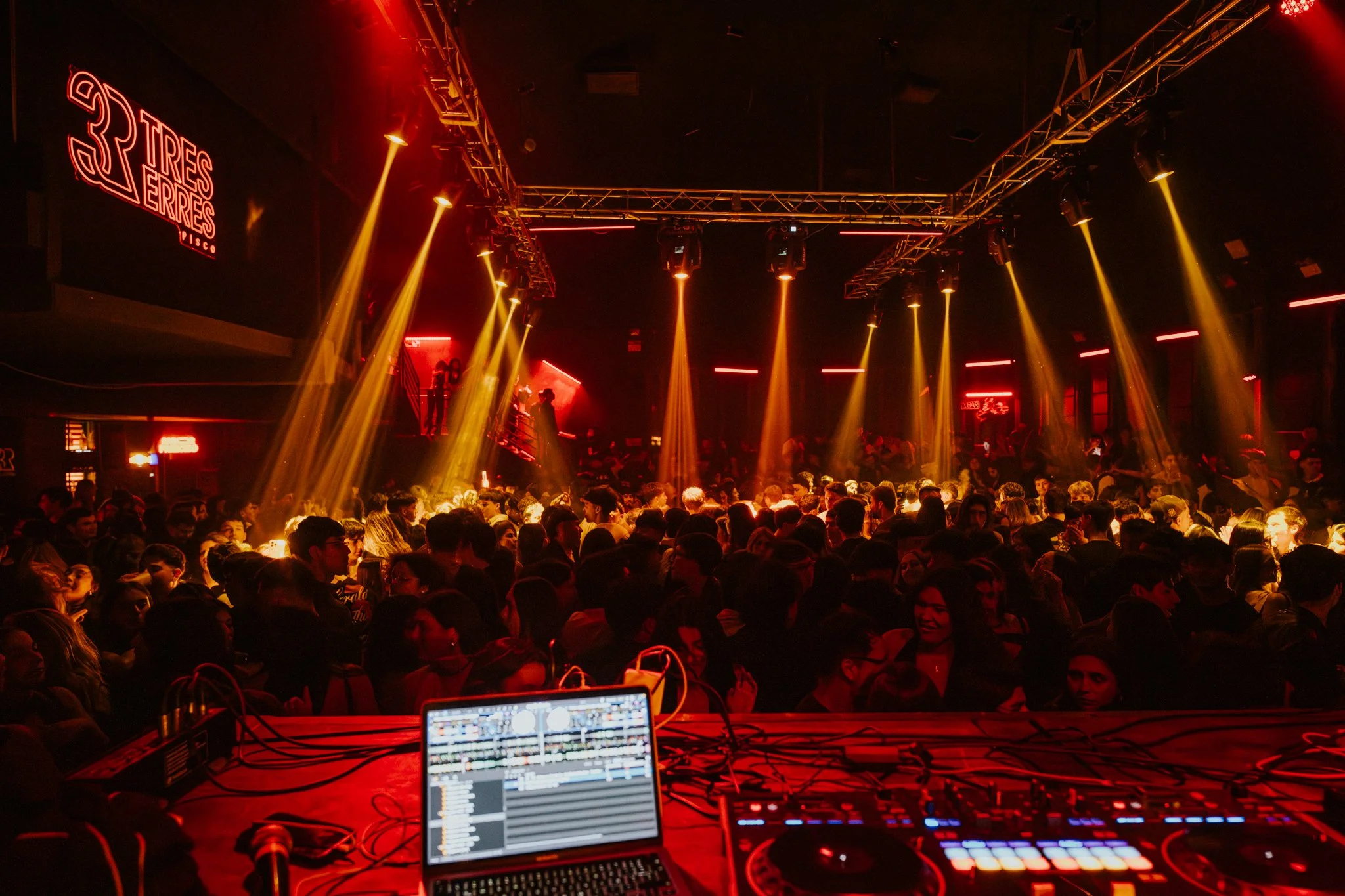 Crowd of people at a nightclub dance floor with colorful, yellow lighting, DJ booth with electronic equipment at the front, and neon signs on dark walls.