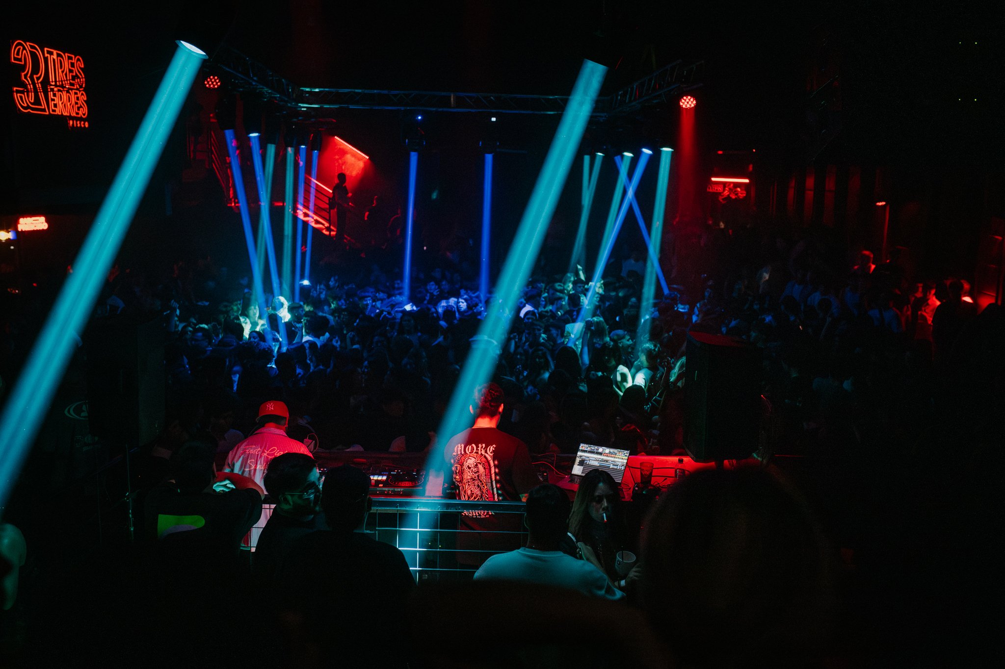 Crowd dancing under colorful stage lights at a nightclub with DJ in the foreground.