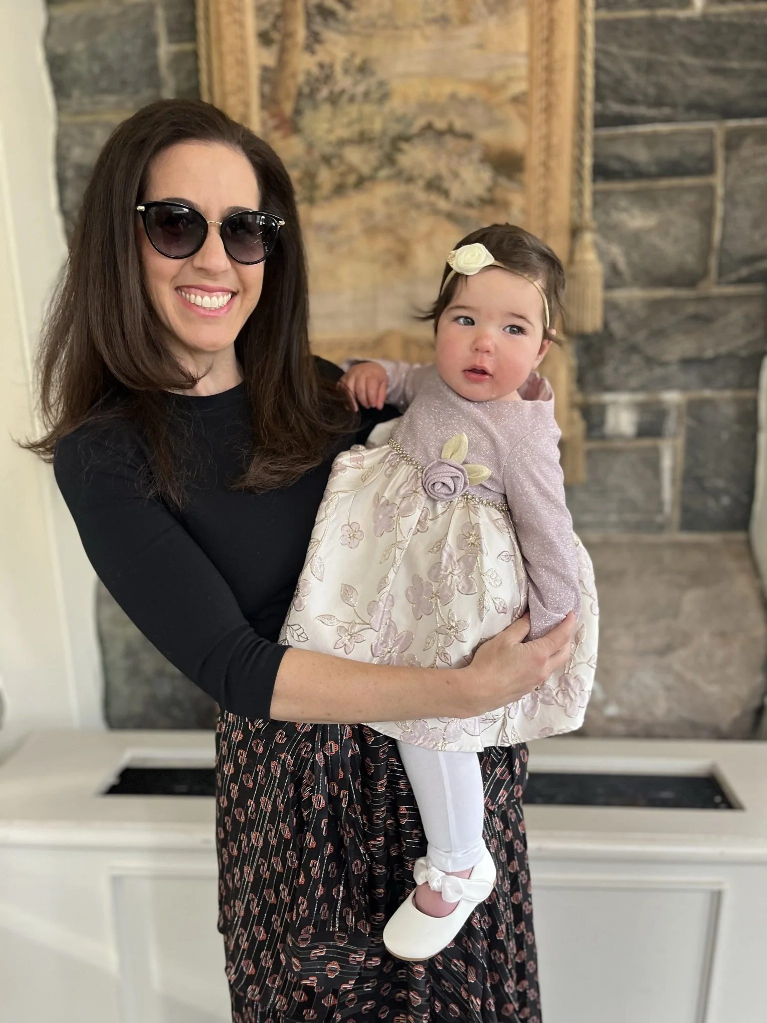 A stylish woman with long brown hair wearing large dark sunglasses and a black top, holding a baby girl dressed in a fancy pink and cream dress with floral details and white shoes, standing in a room with a stone fireplace in the background.