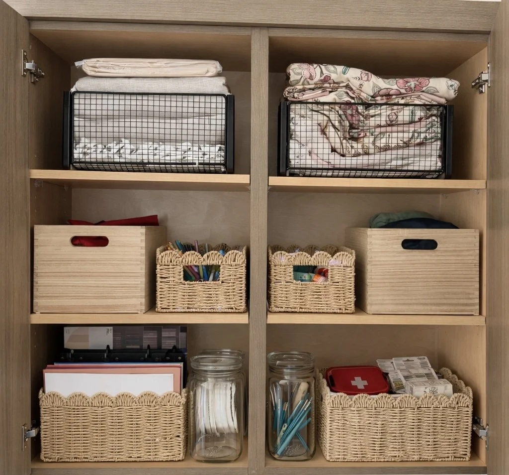 A wooden cabinet containing neatly folded bedsheets, blankets, and pillowcases in wire baskets on top. The middle shelves hold wooden and wicker storage boxes with assorted items like stationery, first aid supplies, and documents. The bottom shelves have wicker baskets with files, glass jars with cotton pads and cotton swabs, and a red first aid kit.