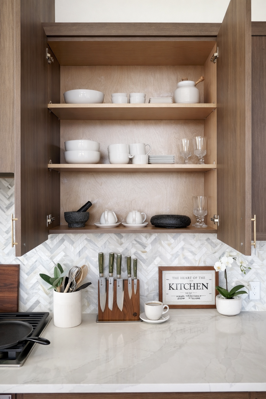 Open kitchen cabinet with white dishes and glasses, a wooden knife block, a white sign reading 'The Heart of the Kitchen,' a white vase with a plant, and a cup of coffee on a marble countertop.