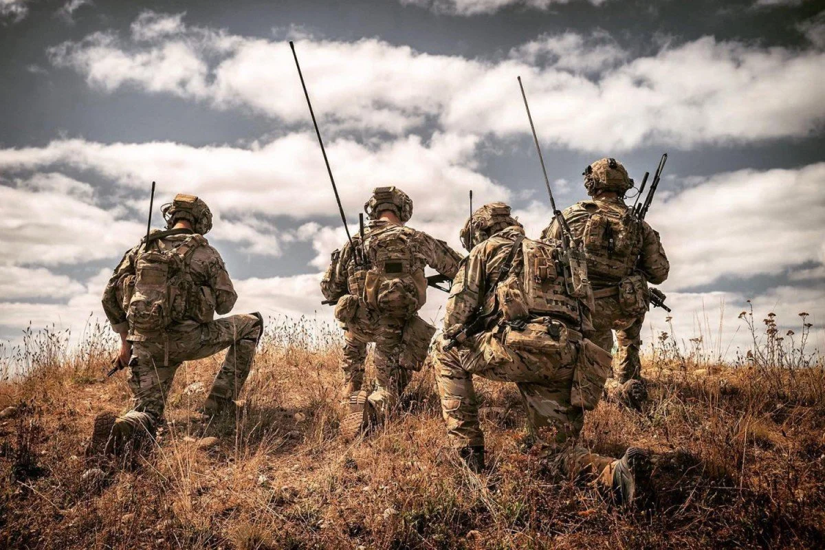 A group of five soldiers in camouflage gear kneeling and standing in a grassy field under a cloudy sky.