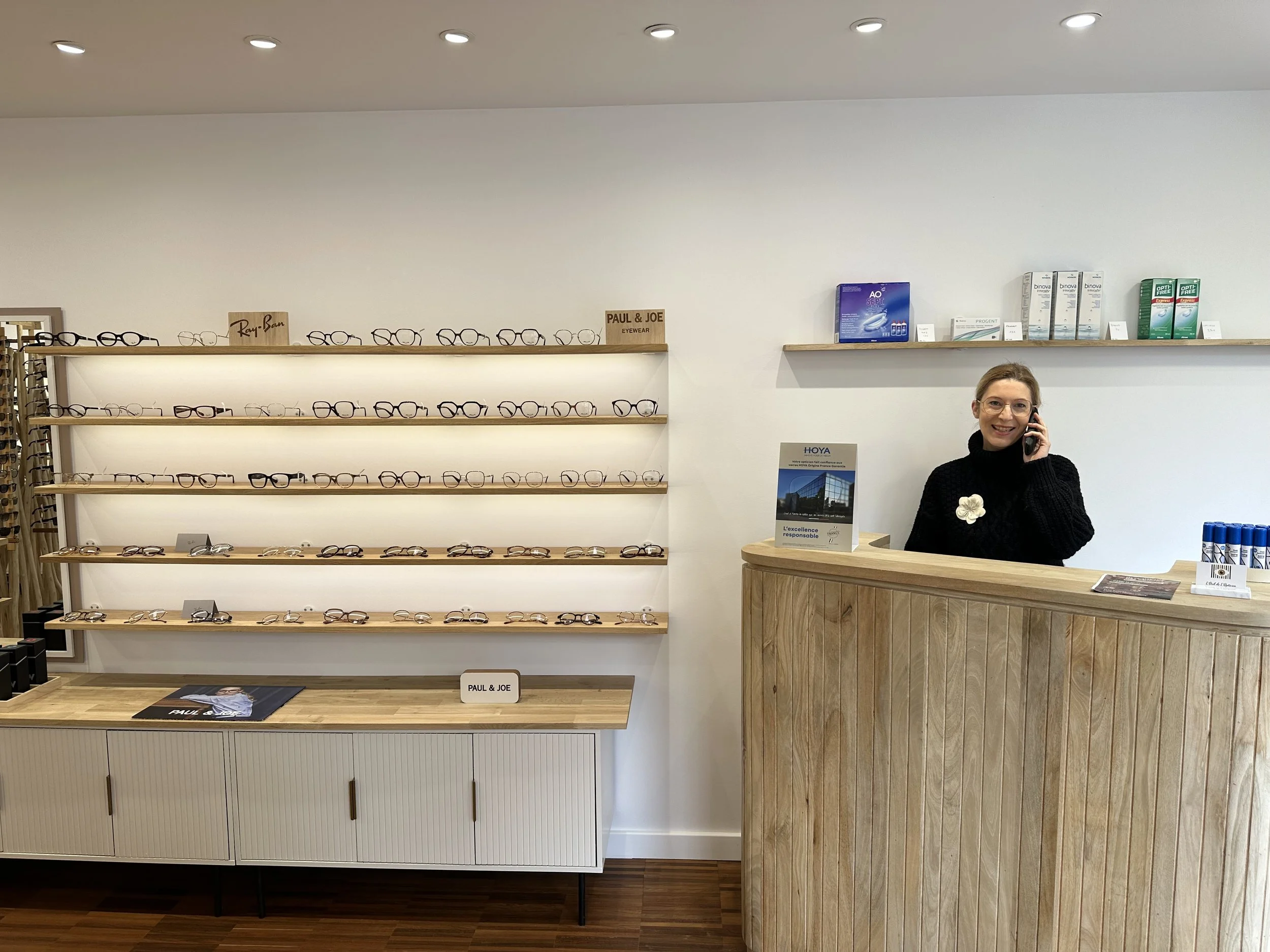 Anne Genay à l'accueil du magasin d'optique L'Oeil de L'Opticien à Bordeaux, avec des étagères de lunettes et des produits pour la vue derrière elle.