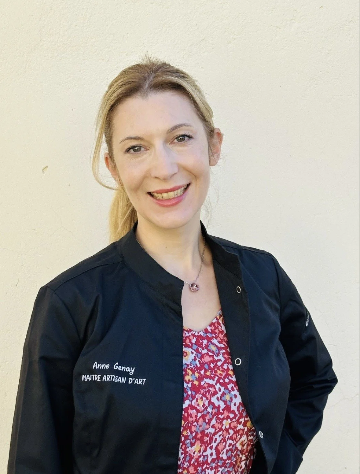 Anne Genay, opticienne, optometriste, maitre artisan d'art,  portant une veste noire avec la mention 'Anne Genay Maître Artisan D'Art' posé sur un fond neutre beige.
