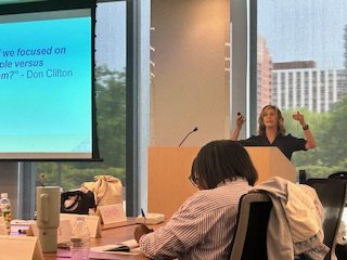 Woman giving a presentation in a conference room with large windows showing a cityscape in the background.