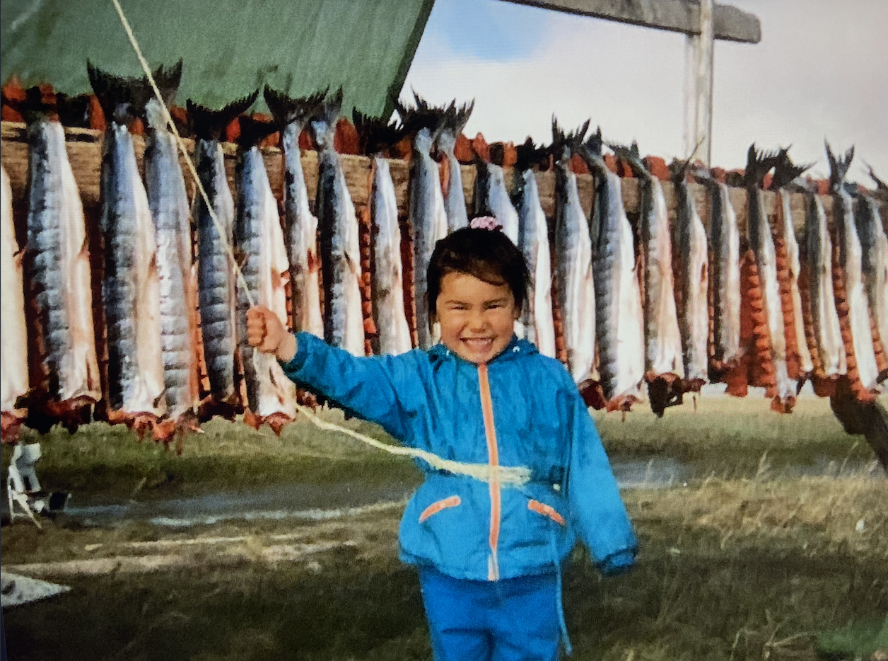 A Cup'ik child at fish camp, standing in front of a rack of fish while holding the tarp from blowing away and smiling at the camera.