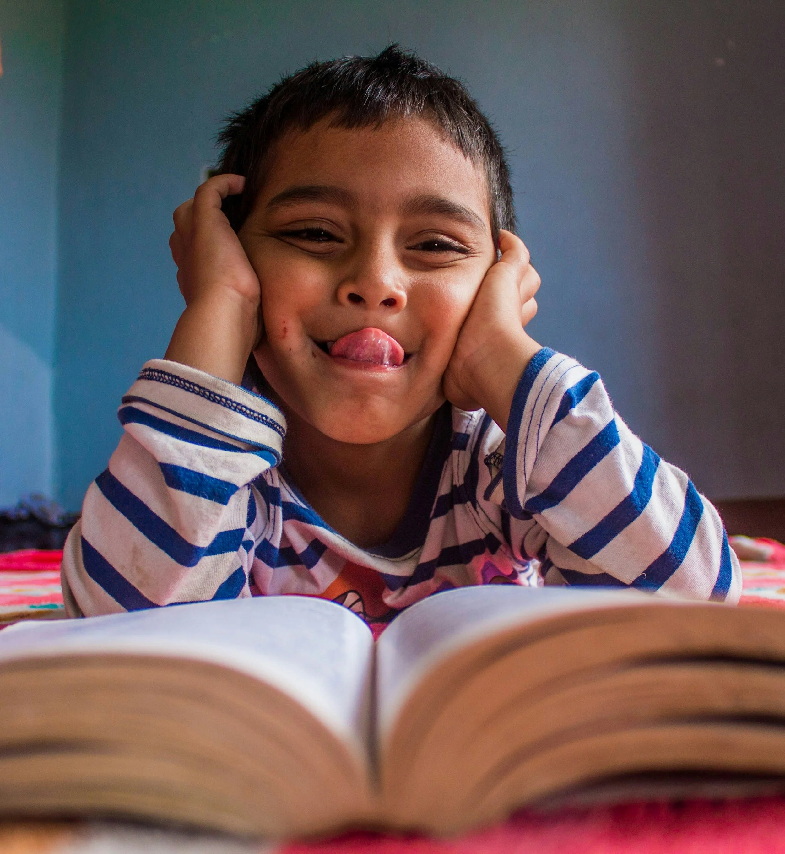 Close-up of a smiling young boy with short dark hair, head resting in hands, wearing a striped blue and white shirt, playful expression sticking tongue out, indoors with a colorful blanket visible.