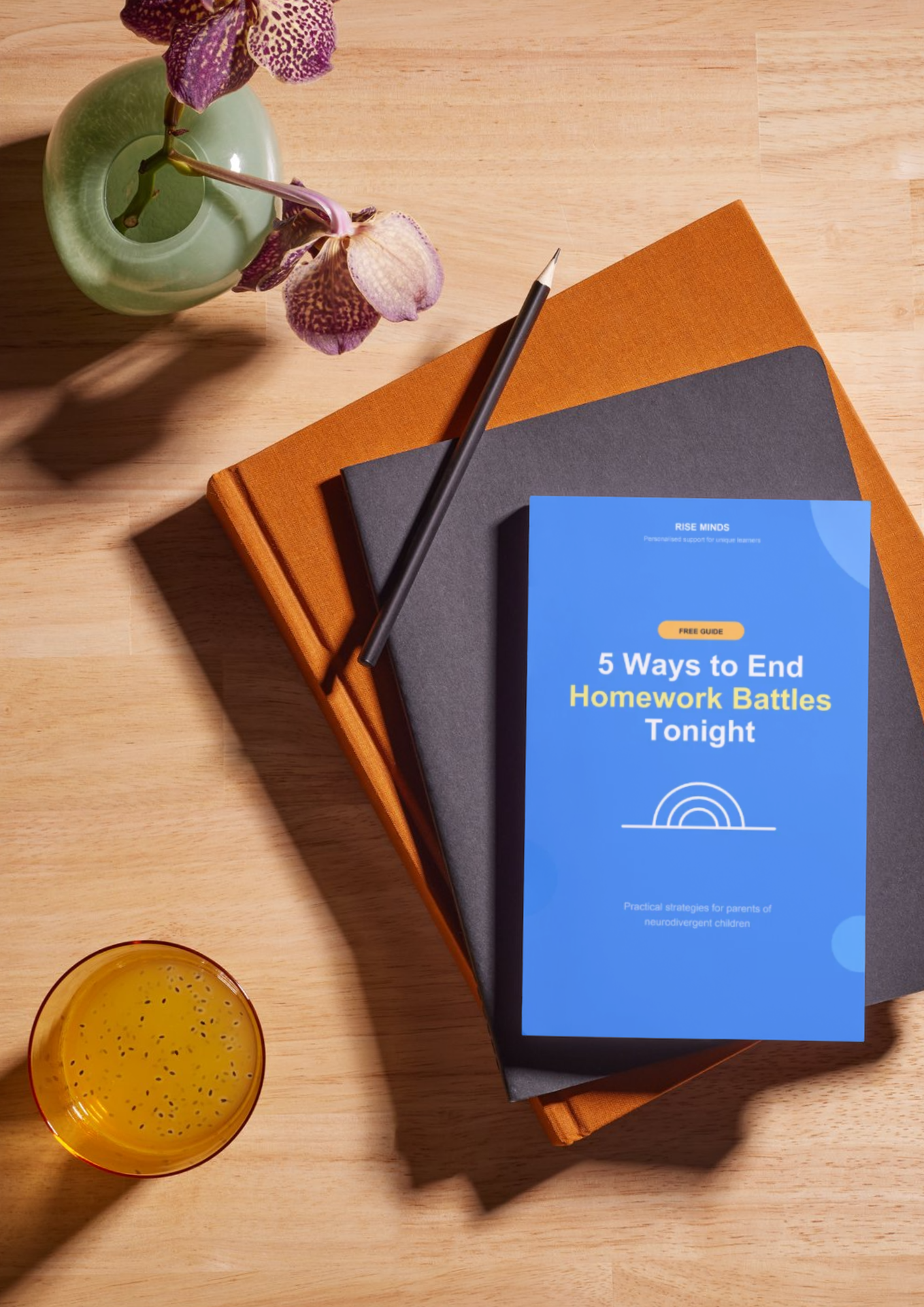 A wooden table with a vase of purple orchids, a small glass of yellow beverage with black seeds, and two books, one orange and one black, with a blue booklet titled "5 Ways to End Homework Battles Tonight".
