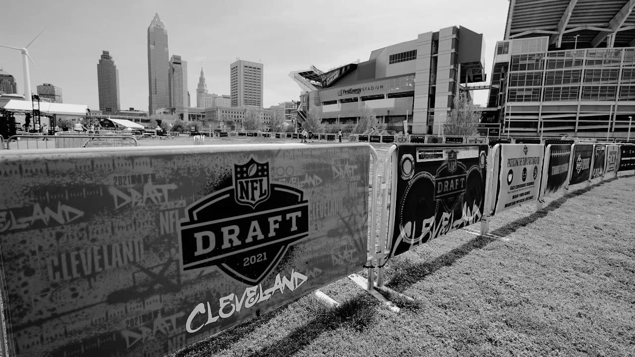 Black and white photo of NFL Draft signage along a sidewalk at FirstEnergy Stadium in Cleveland, with city skyline including tall buildings in the background.