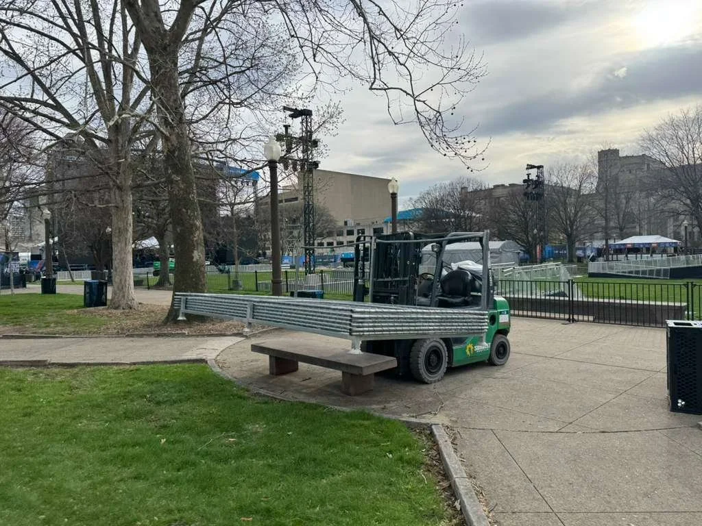 Empty park with trees, a bench, a forklift, and a fenced-off area with tents in the background.