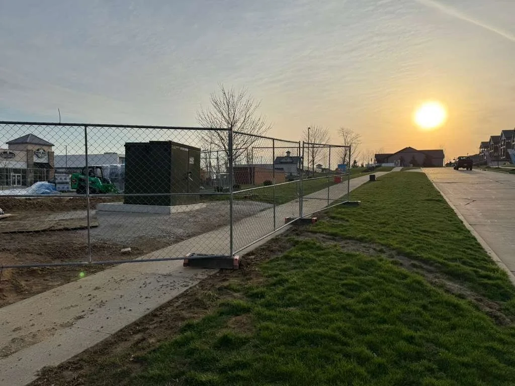 Sunset over a residential neighborhood with a sidewalk and new grass, a construction fence, and a utility box in the foreground.