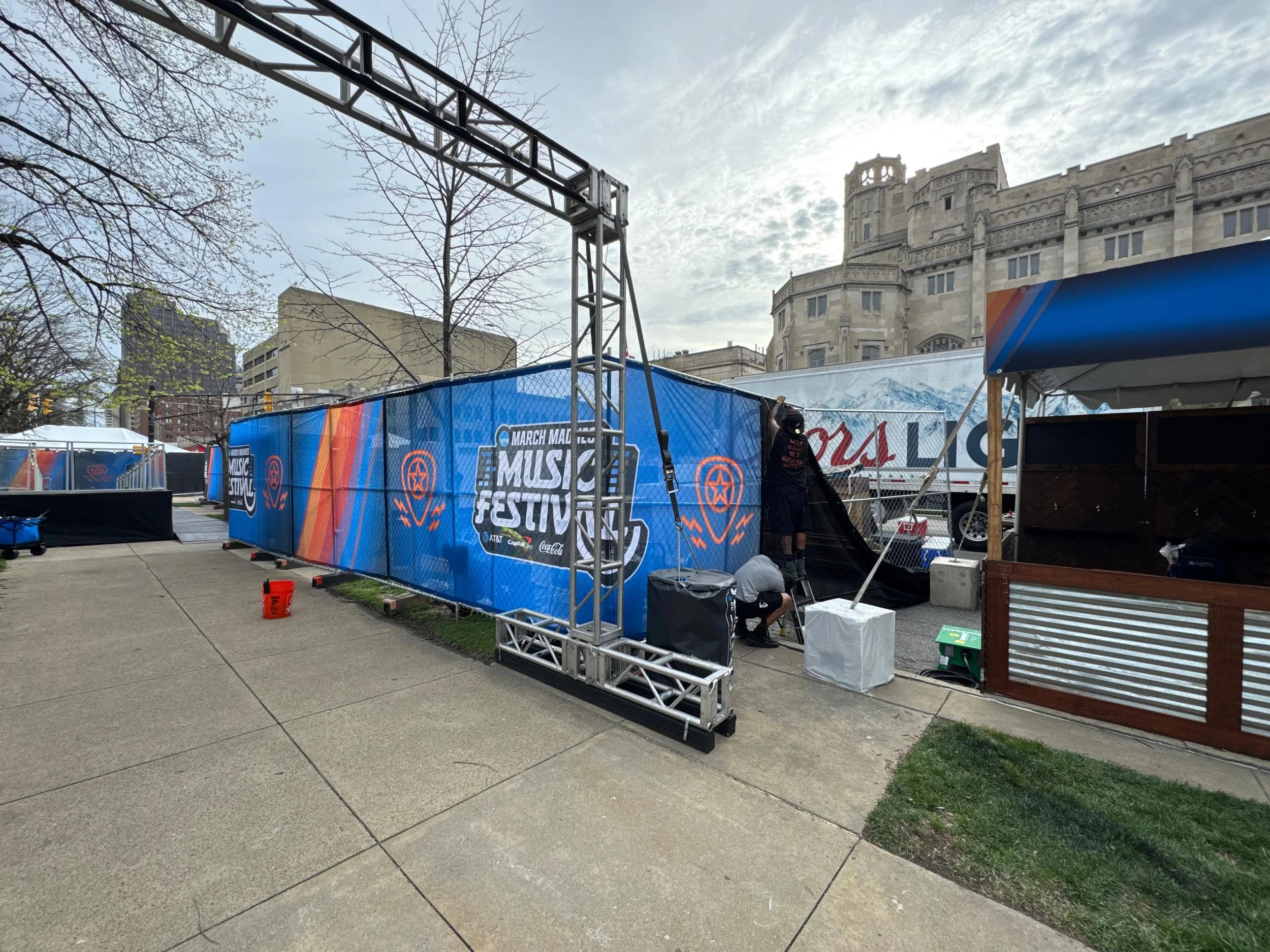 Outdoor scene of a music festival setup in a city park, with a blue barricade fence that reads 'March Music Festival', a stage being assembled, and historic buildings in the background.