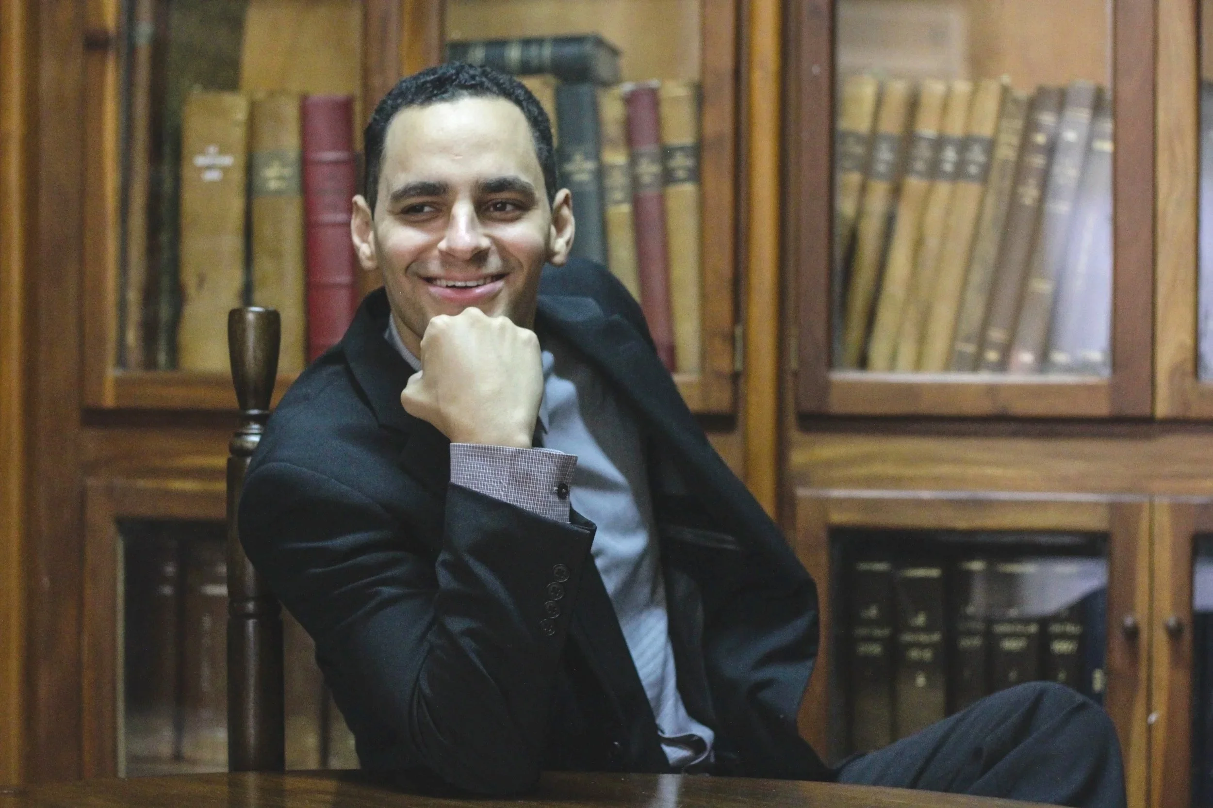 Alexandre de Lespinasse, 5th generation owner, in a black suit sitting in a wooden library with glass cabinets filled with old books behind him, smiling with his chin resting on his hand.