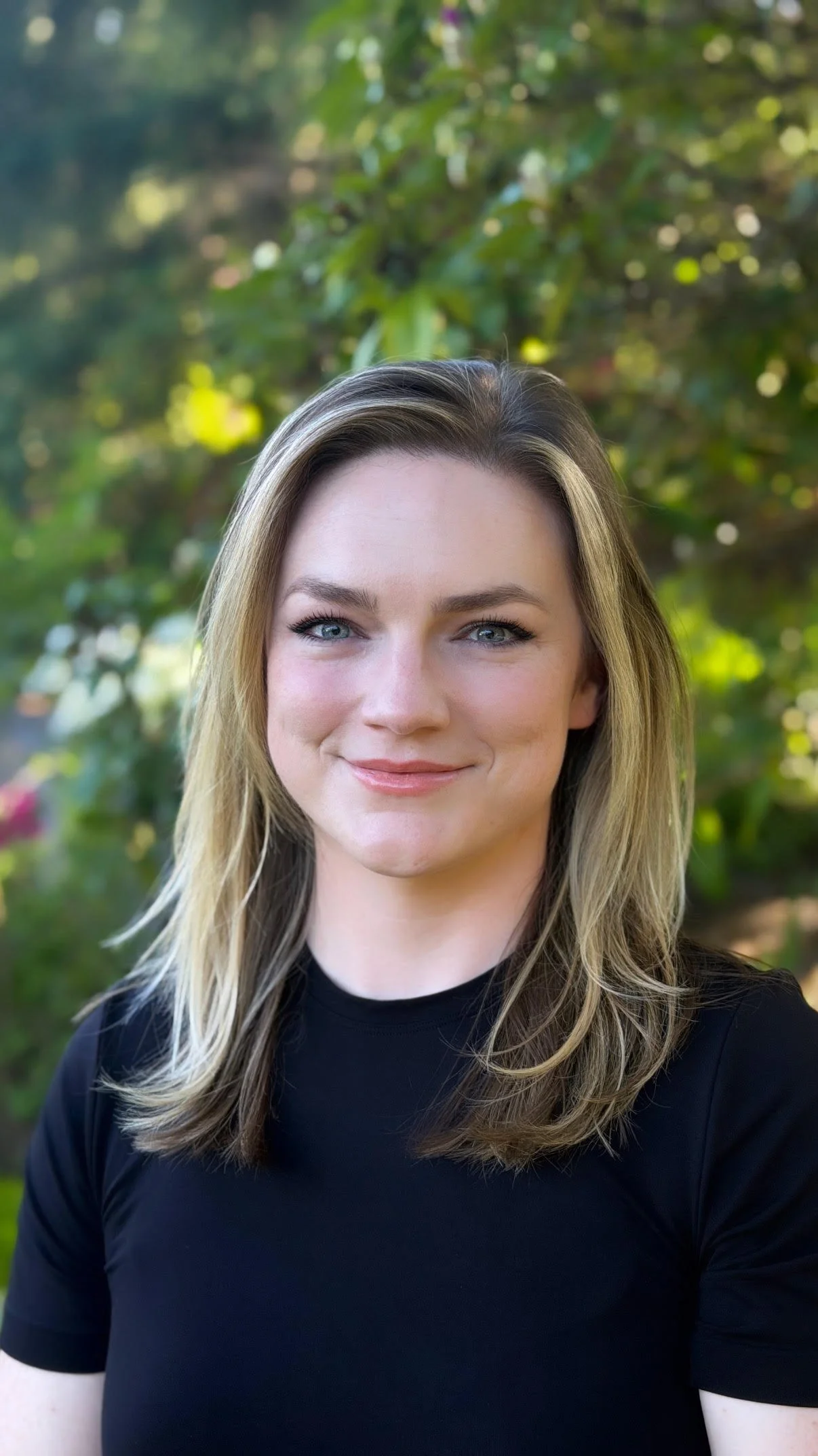A young woman with blonde hair and blue eyes smiling outdoors with a background of greenery.