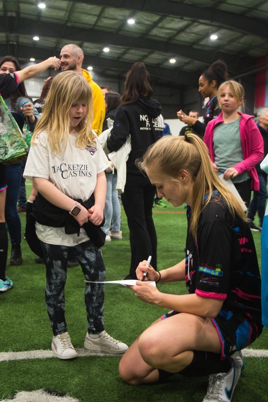A woman is kneeling on one knee on a green indoor turf field, signing an autograph on a piece of paper with a marker. A young girl with long blonde hair and a MacKenzie Castings t-shirt stands in front of her, watching. Several other children and adults are gathered in the background inside a large indoor sports facility with a curved ceiling and bright lighting.