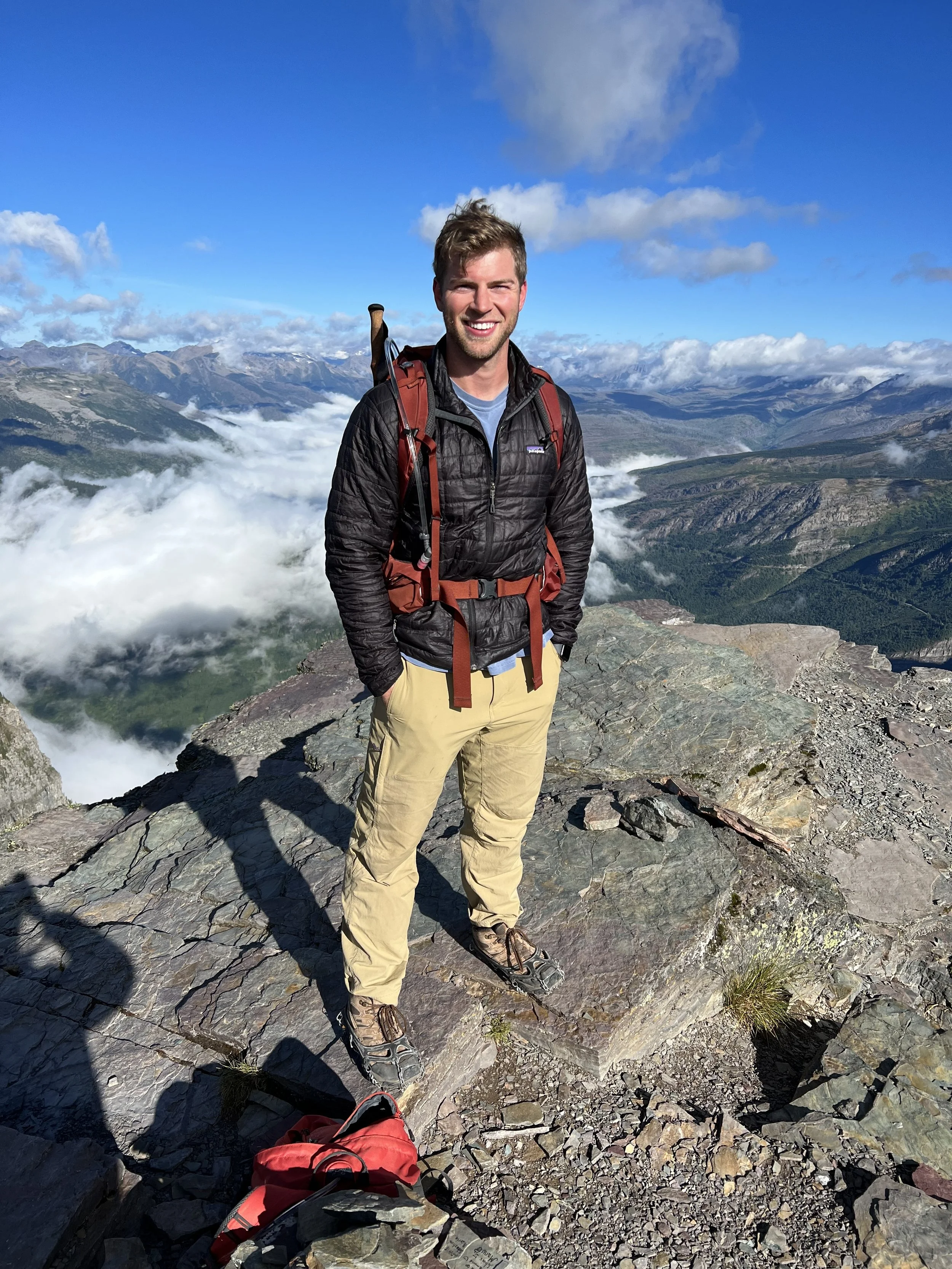 A smiling man with a backpack standing on a rocky ledge in a mountainous landscape with clouds, mountains, and blue sky in the background.