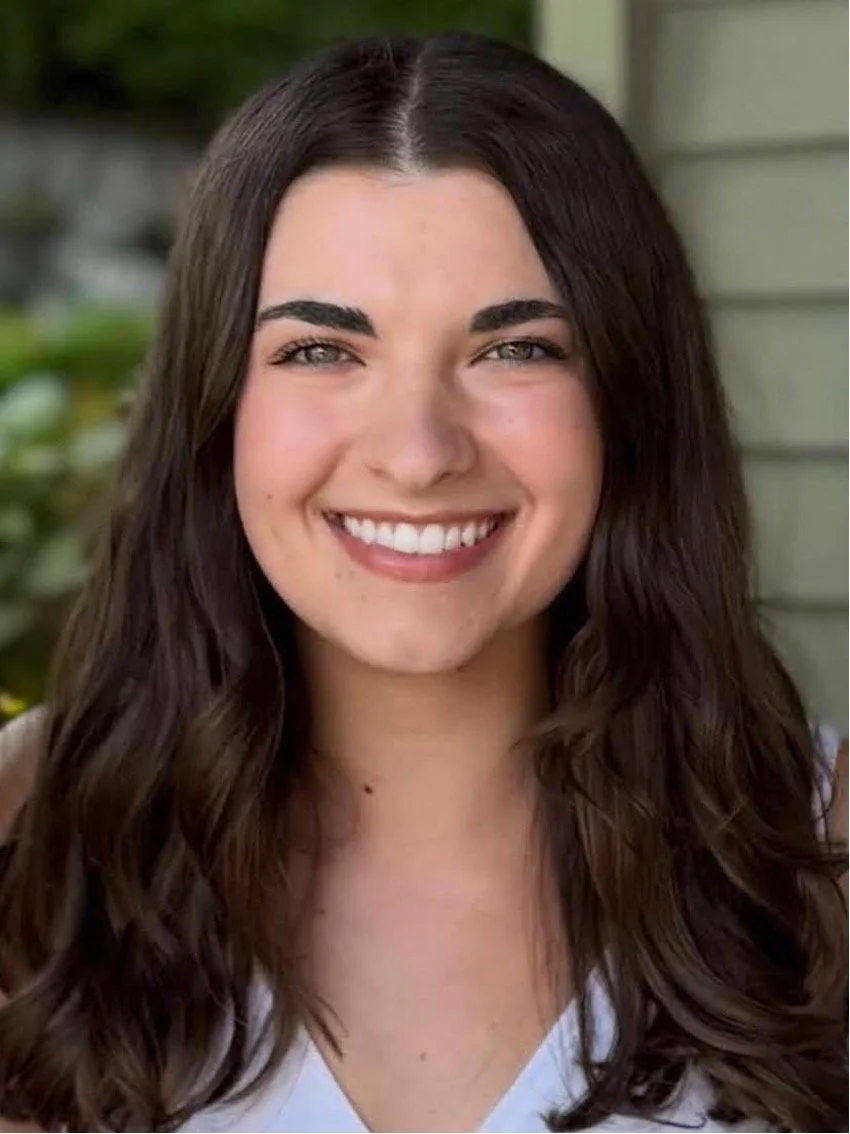 A young woman with long wavy brown hair, smiling showing her teeth, outdoors with greenery and house siding in the background.
