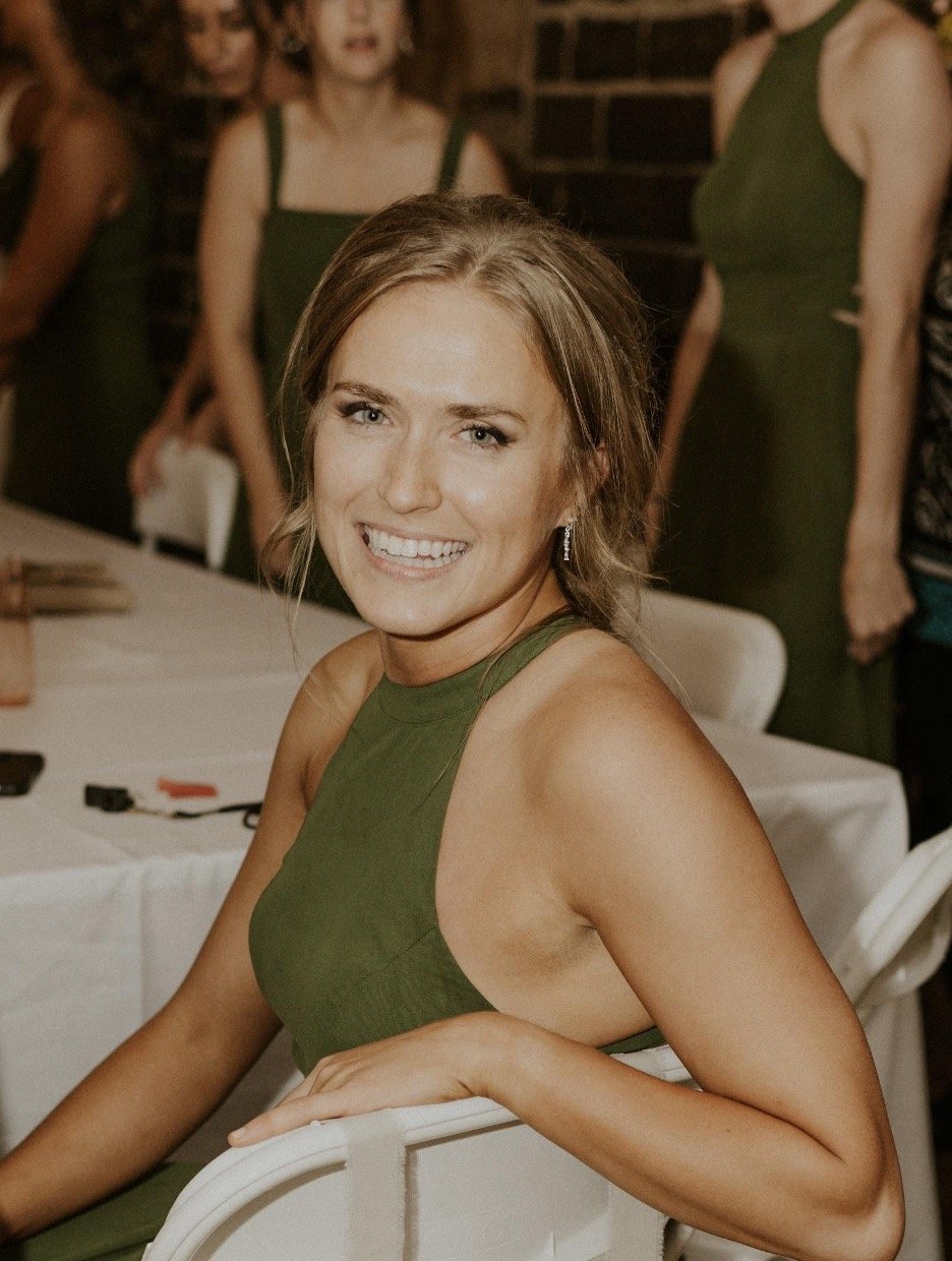 A smiling woman with light brown hair in a green sleeveless dress, sitting at a table indoors, with other women in green dresses standing behind her.