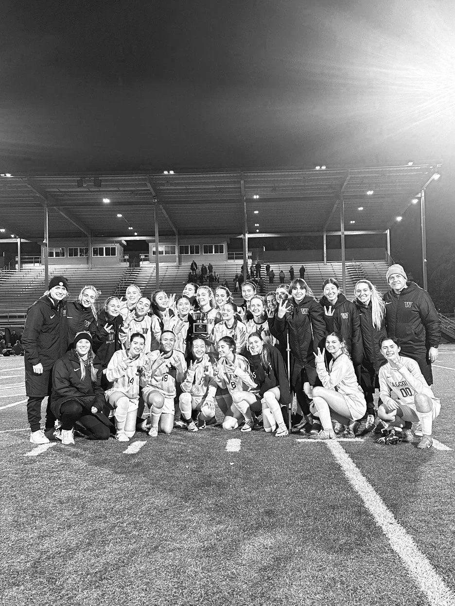 A group of female soccer players and coaches posing on a field at night, some holding a trophy, with stadium stands and lights in the background.