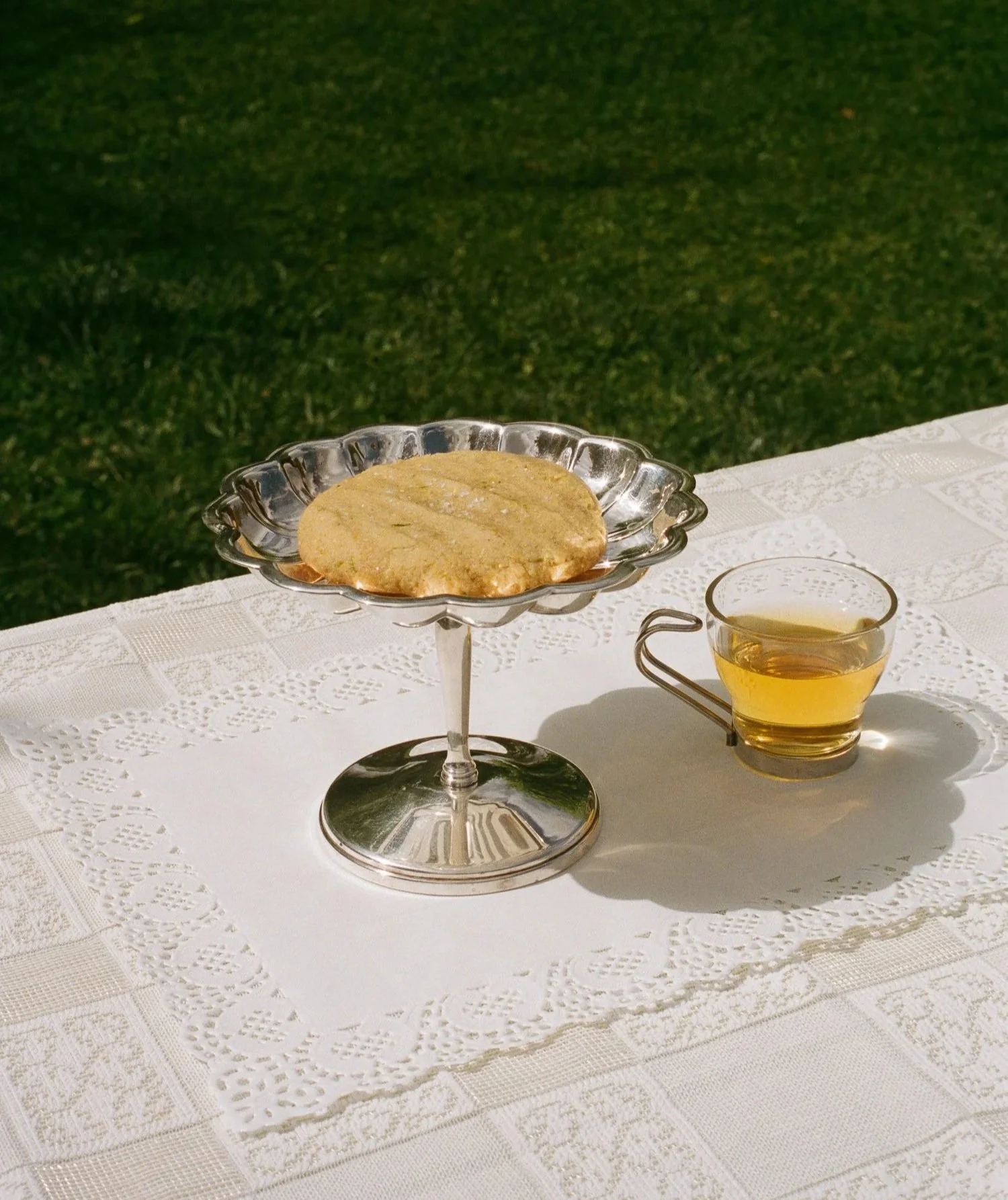 A metal cake stand with a large round cookie on top, next to a small glass cup of tea, all on a white lace tablecloth outdoors with green grass in the background.