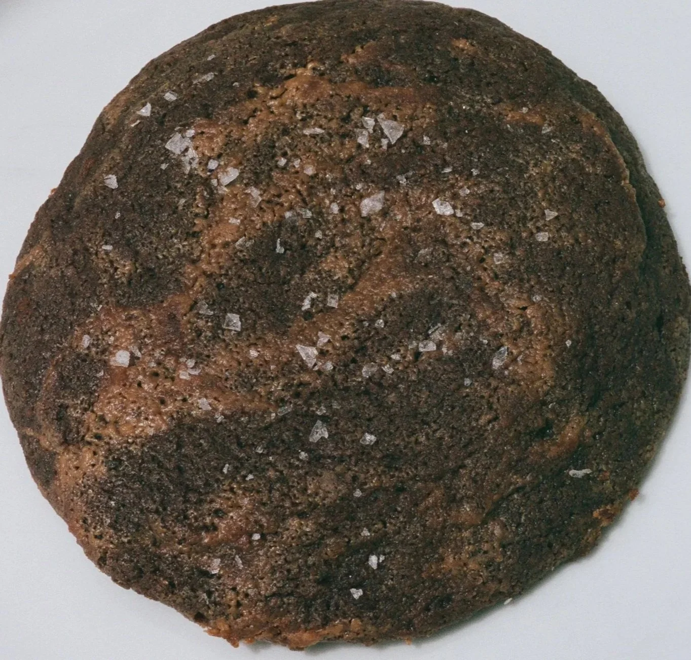 A close-up view of a round, chocolate cookie with a cracked surface, sprinkled with coarse sea salt flakes.