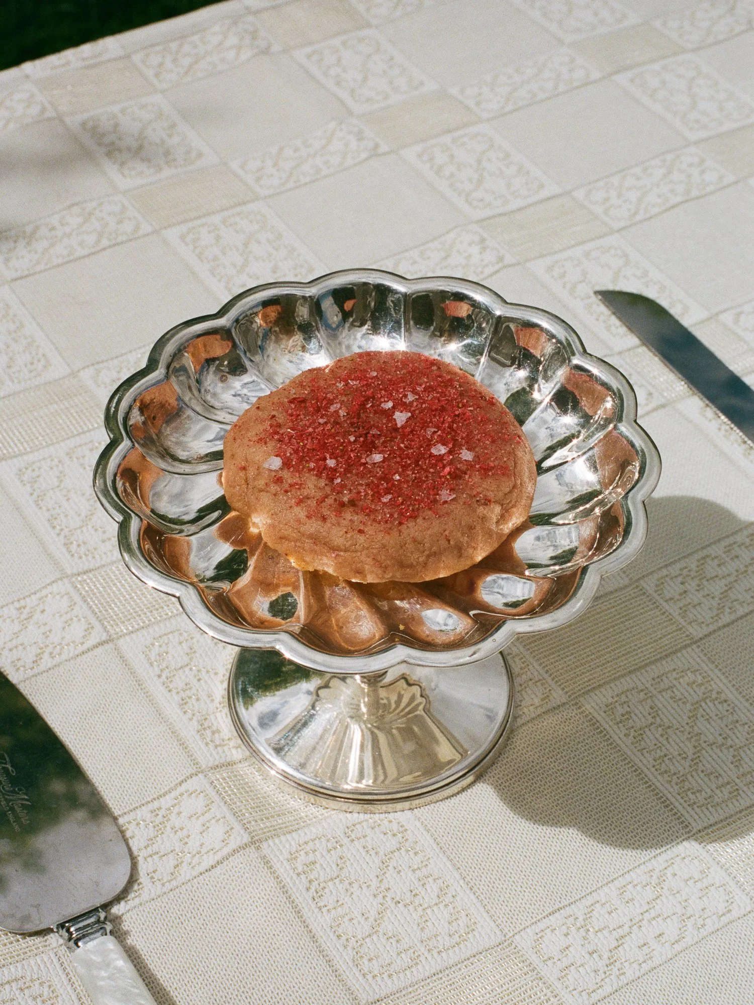 A dessert cookie covered with red sprinkles on a decorative silver dish, placed on a white textured tablecloth with a knife nearby.