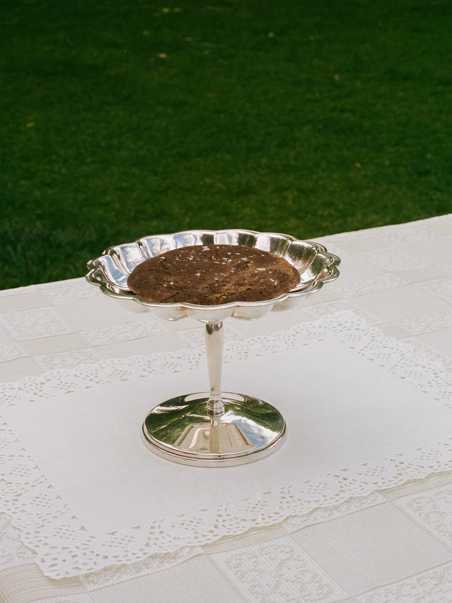 A chocolate cookie on a silver decorative cake stand set on a white lace tablecloth outside with green grass in the background.