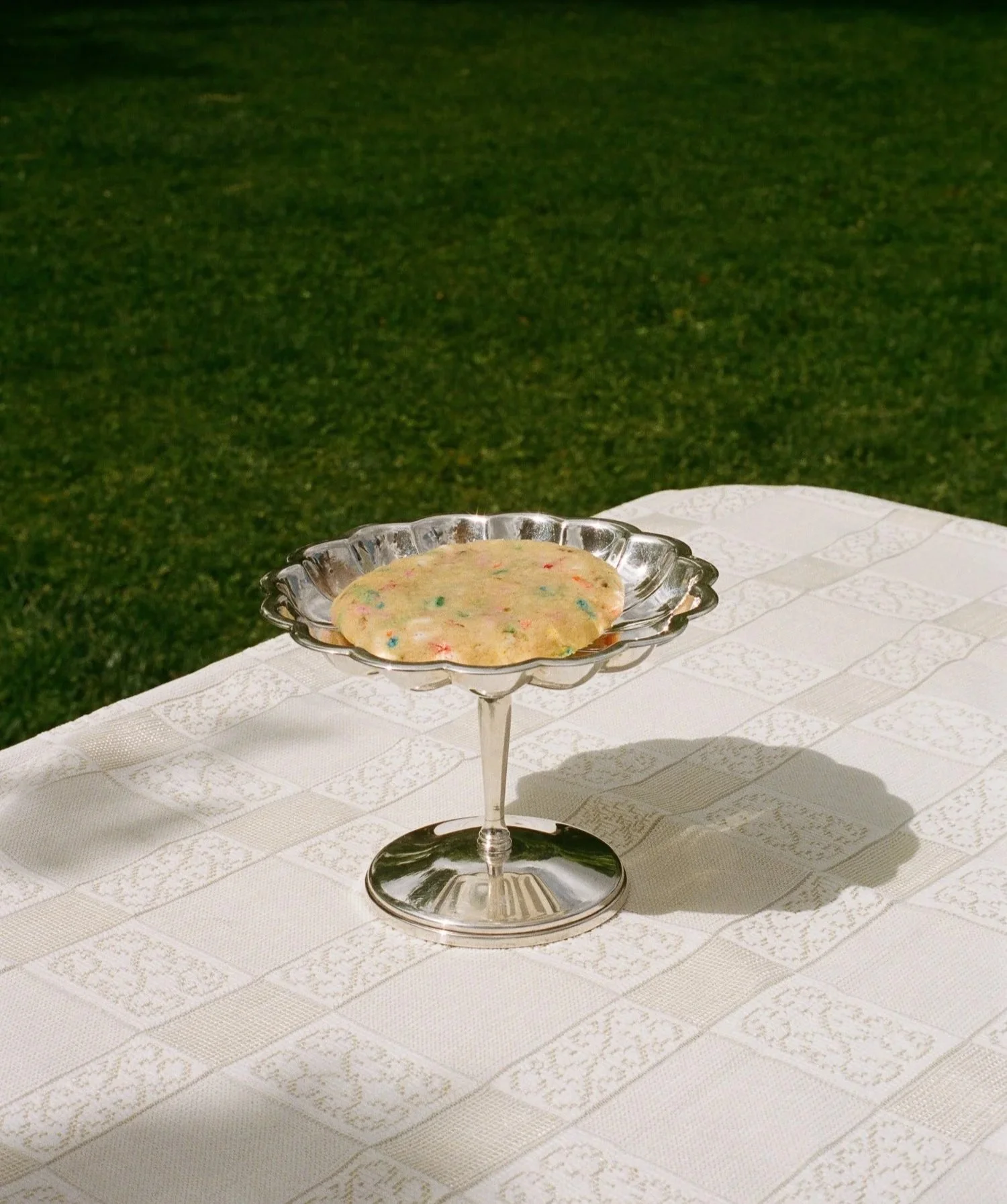 A glass dessert stand with a colorful cookie on top, placed on a white patterned tablecloth outdoors with green grass in the background.