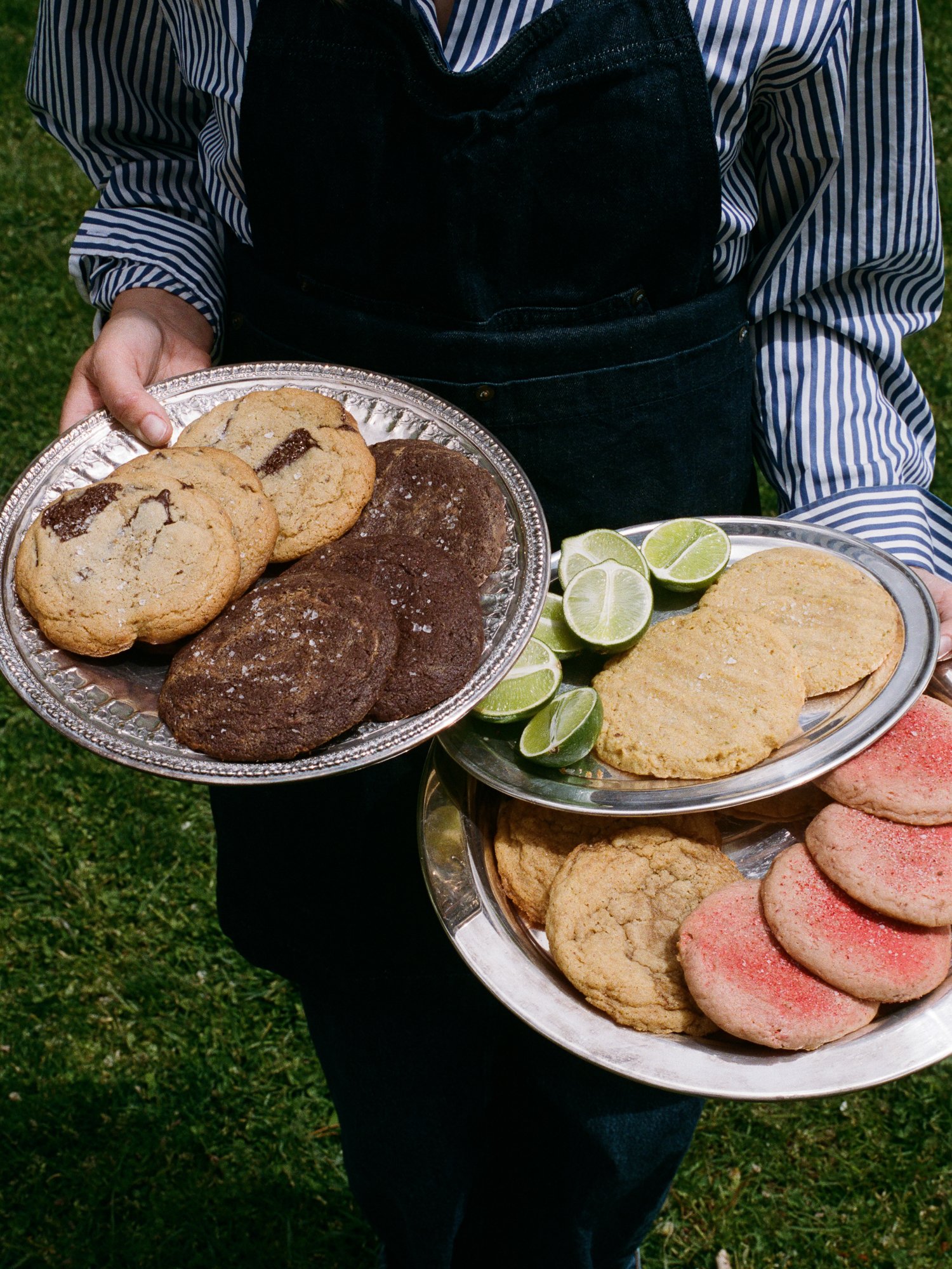 Person holding two silver platters of assorted cookies and lime wedges, wearing a striped shirt and dark apron outdoors.
