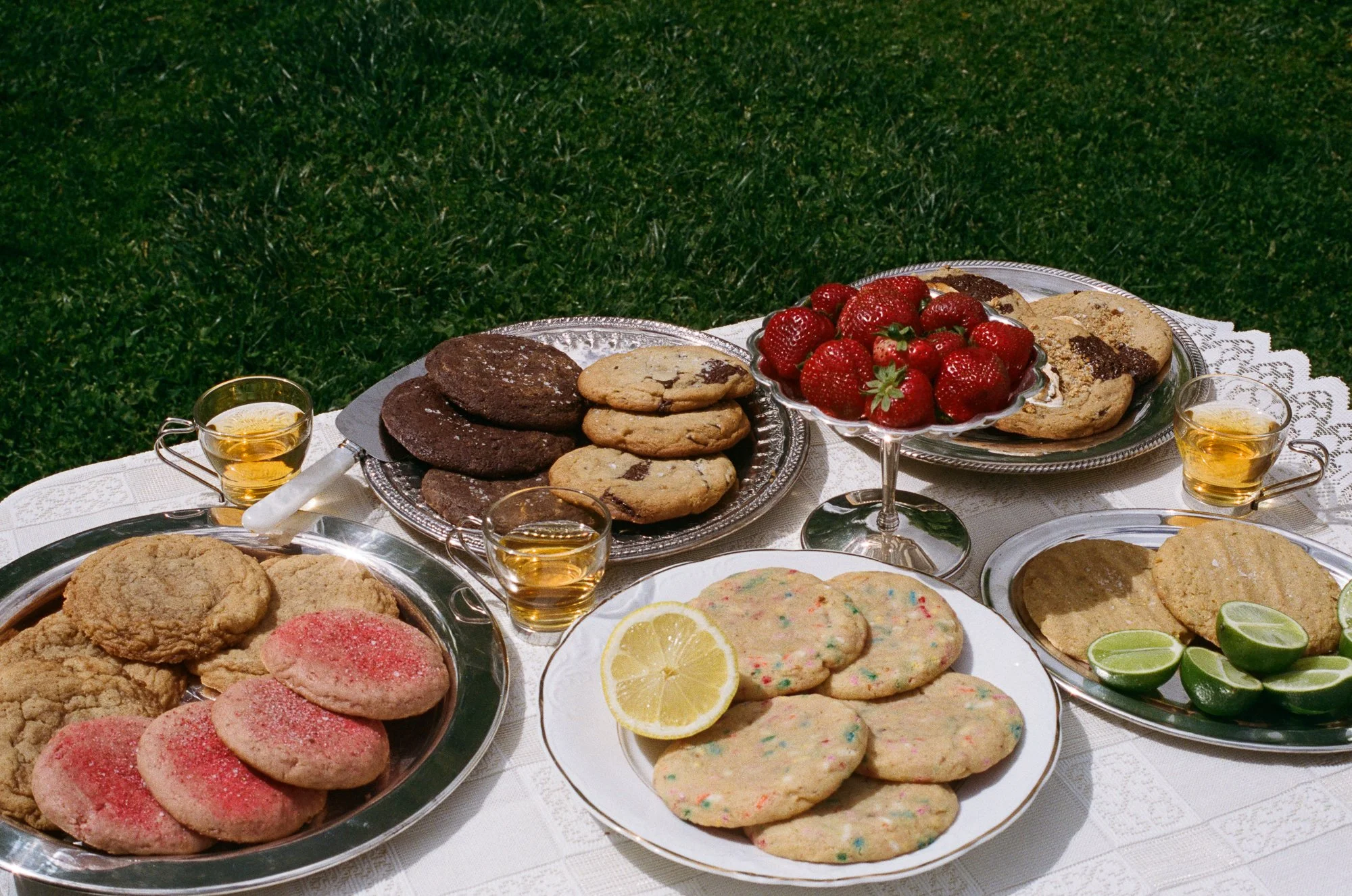 lace tablecloth with silver trays of cookies piled high. chocolate chip, strawberry, funfetti, cornbread, smores, snickerdoodle