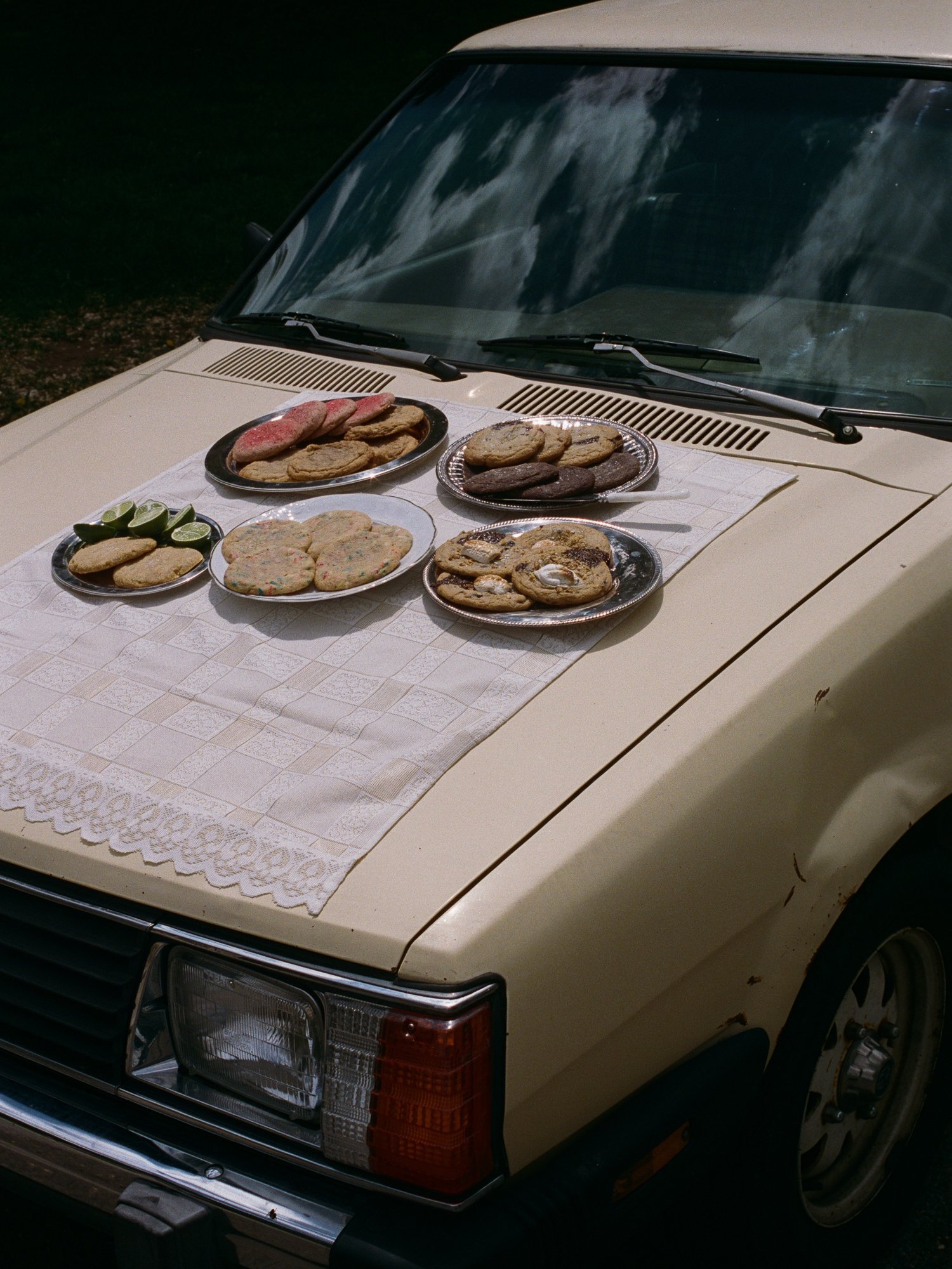 Cookies and lime wedges placed on the hood of an old beige car, which has a white lace cloth on the hood, reflecting blue sky and clouds in the windshield.