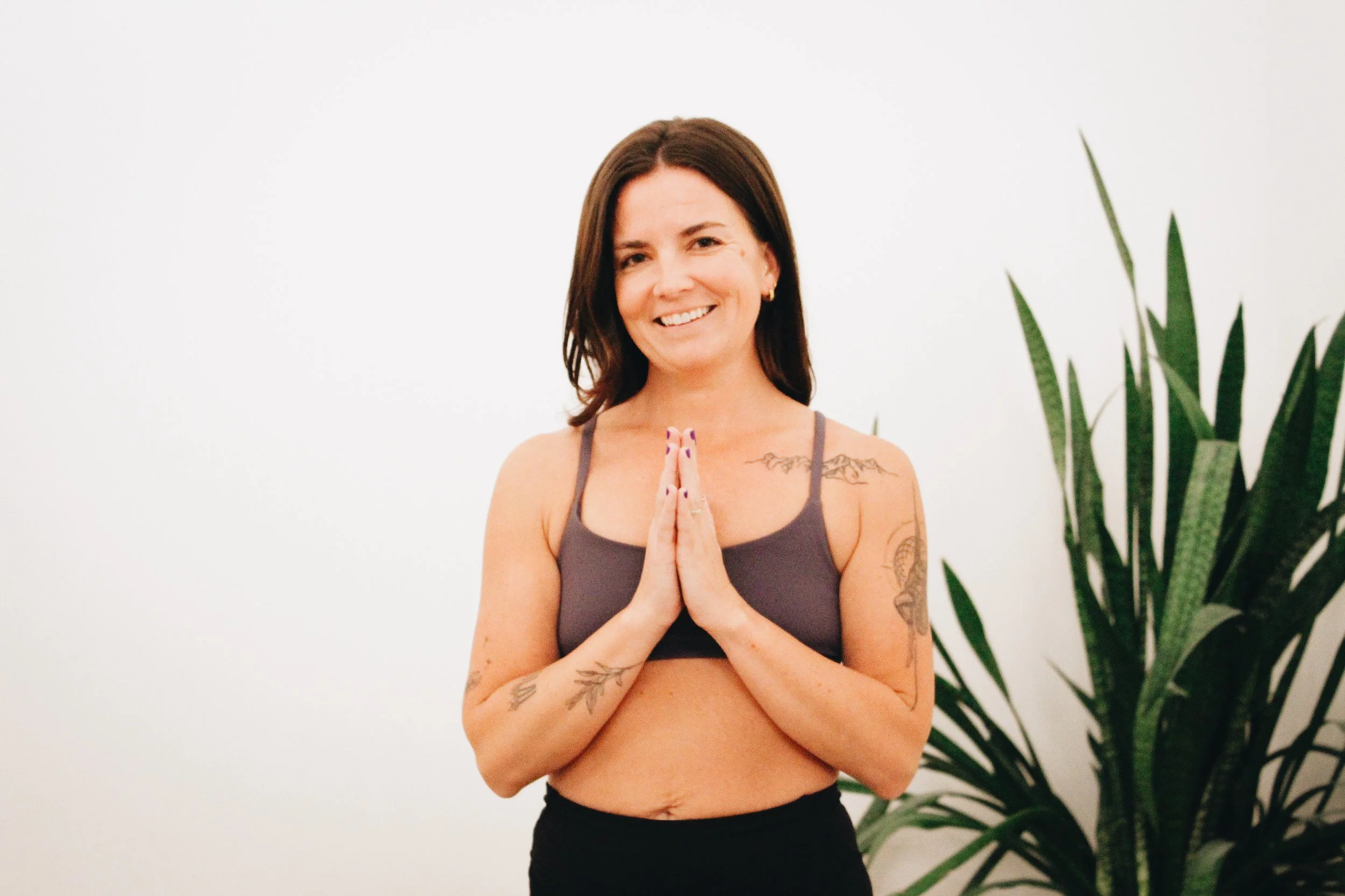 A woman smiling, standing with hands in a prayer position, wearing a sports bra and black pants, near a large green plant against a plain white wall.