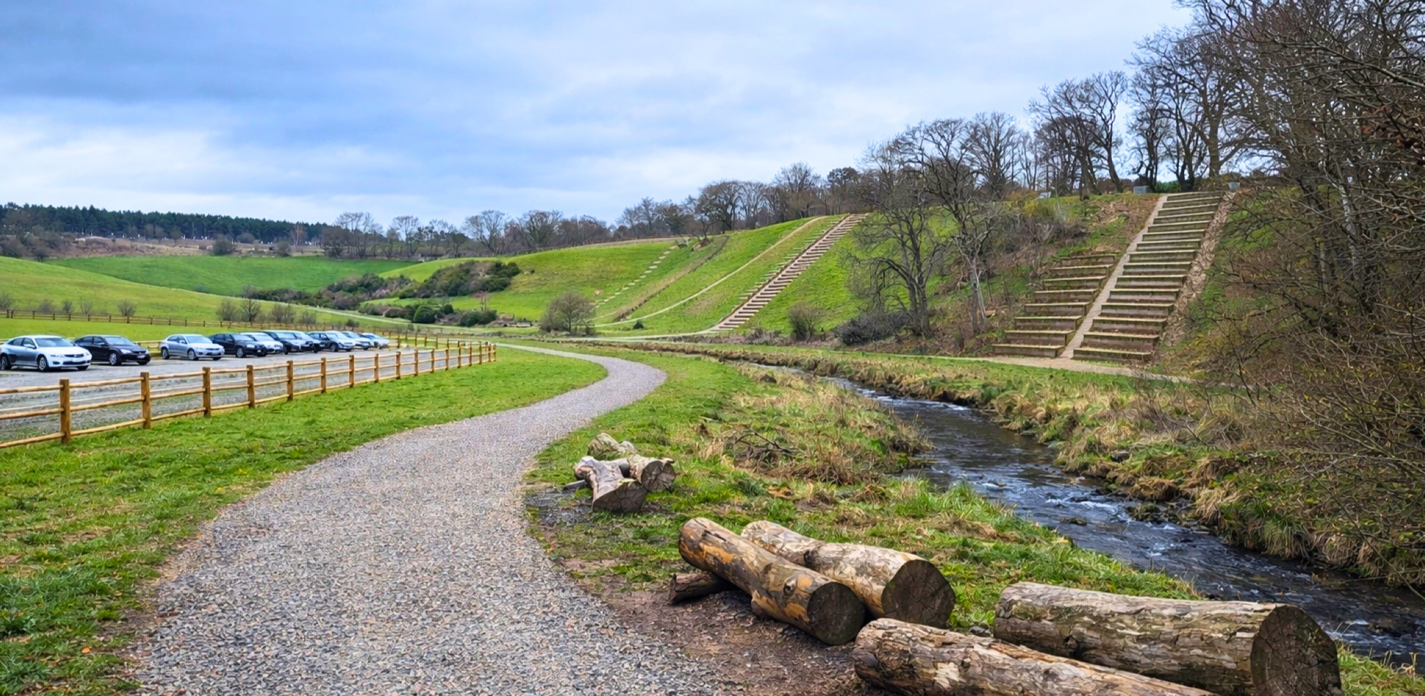 A scenic landscape featuring a gravel path alongside a small stream, with a parking lot filled with cars on the left. In the background, there are green grassy hills with steps and stairs leading up the hillside, and bare trees under a cloudy sky.